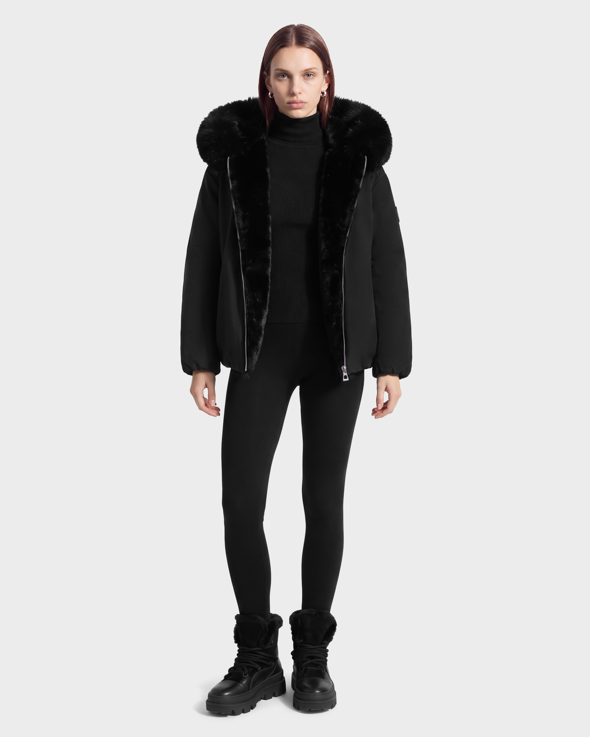 A person wearing a KATHLEEN - 8126764 Black puffer jacket with a fur-trimmed hood, a black turtleneck, black leggings, and black boots.