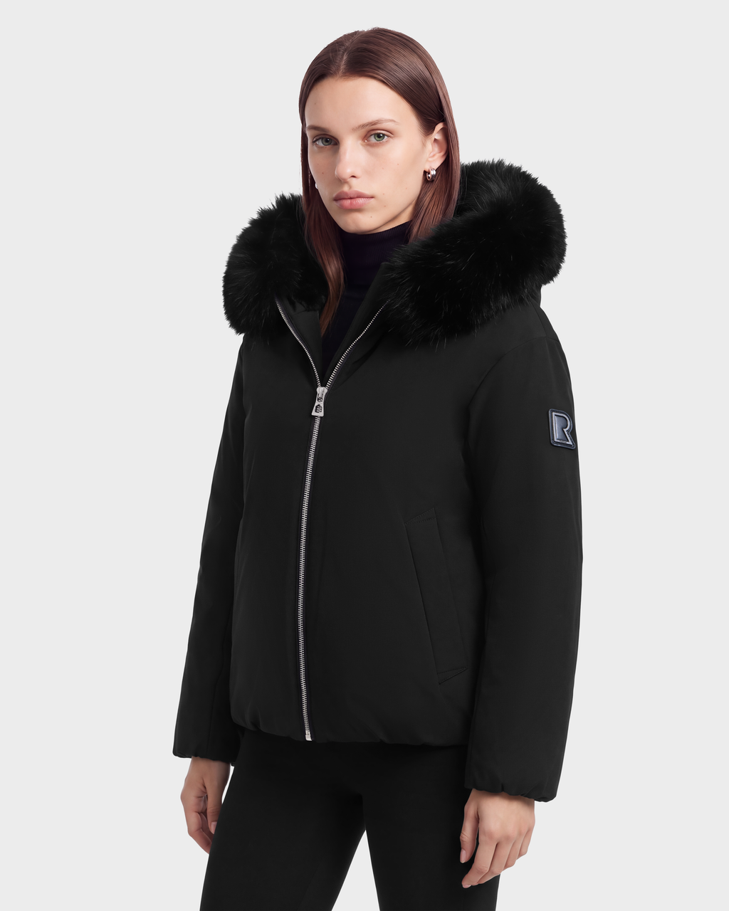 A person wearing a black Rudsak KATHLEEN - 8126764 puffer jacket with a fur-trimmed hood and a blue logo patch on the sleeve.