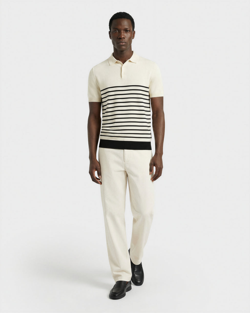 A man wearing Rudsak’s KRIS - 6526005 Iceland cream polo with black stripes and matching cream trousers, paired with black shoes, walks forward against a plain white background.