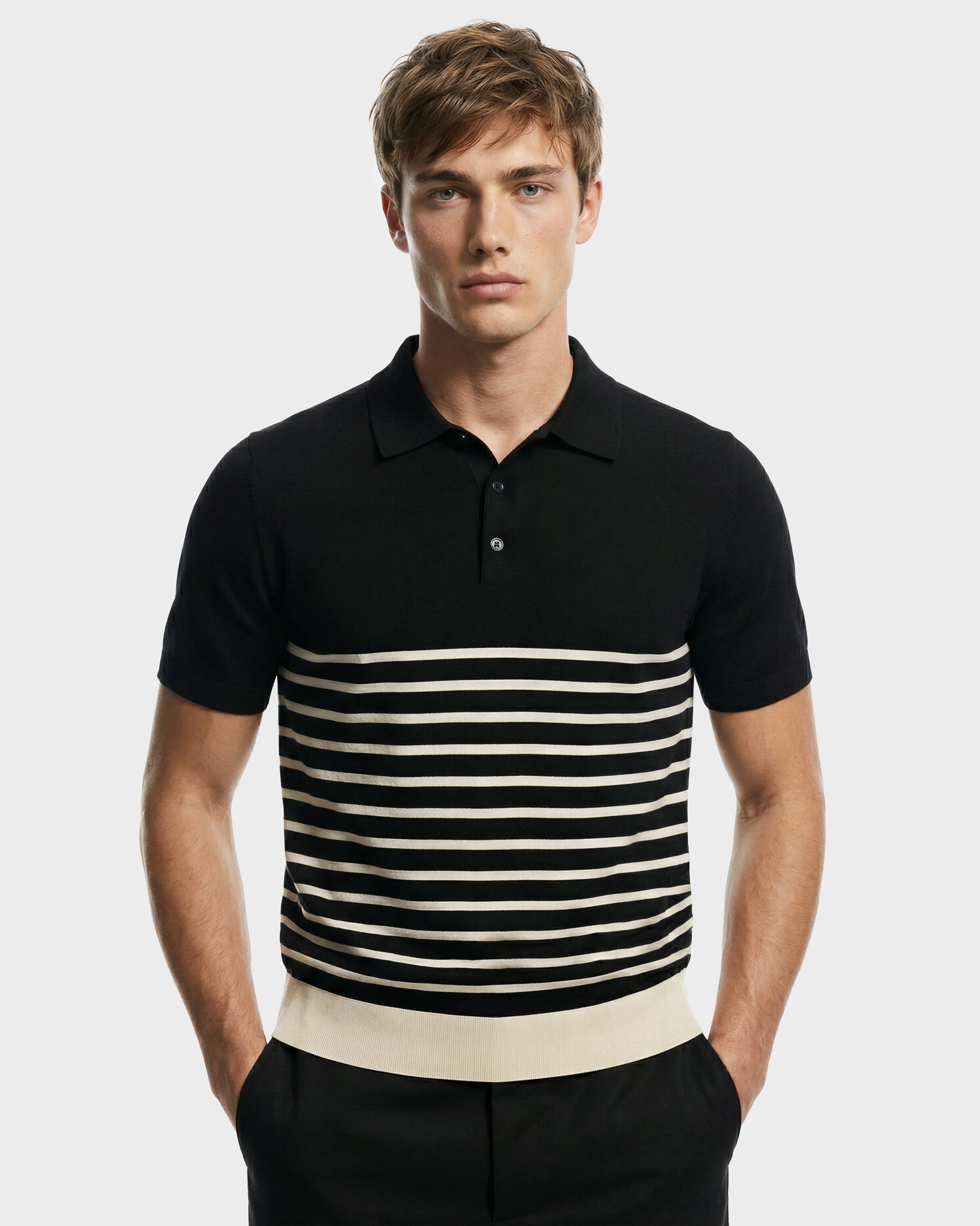 A young man with light brown hair models the KRIS - 6526005 Black men's designer polo by Rudsak, featuring black fabric with white horizontal stripes, standing hands-in-pockets against a plain light gray background.