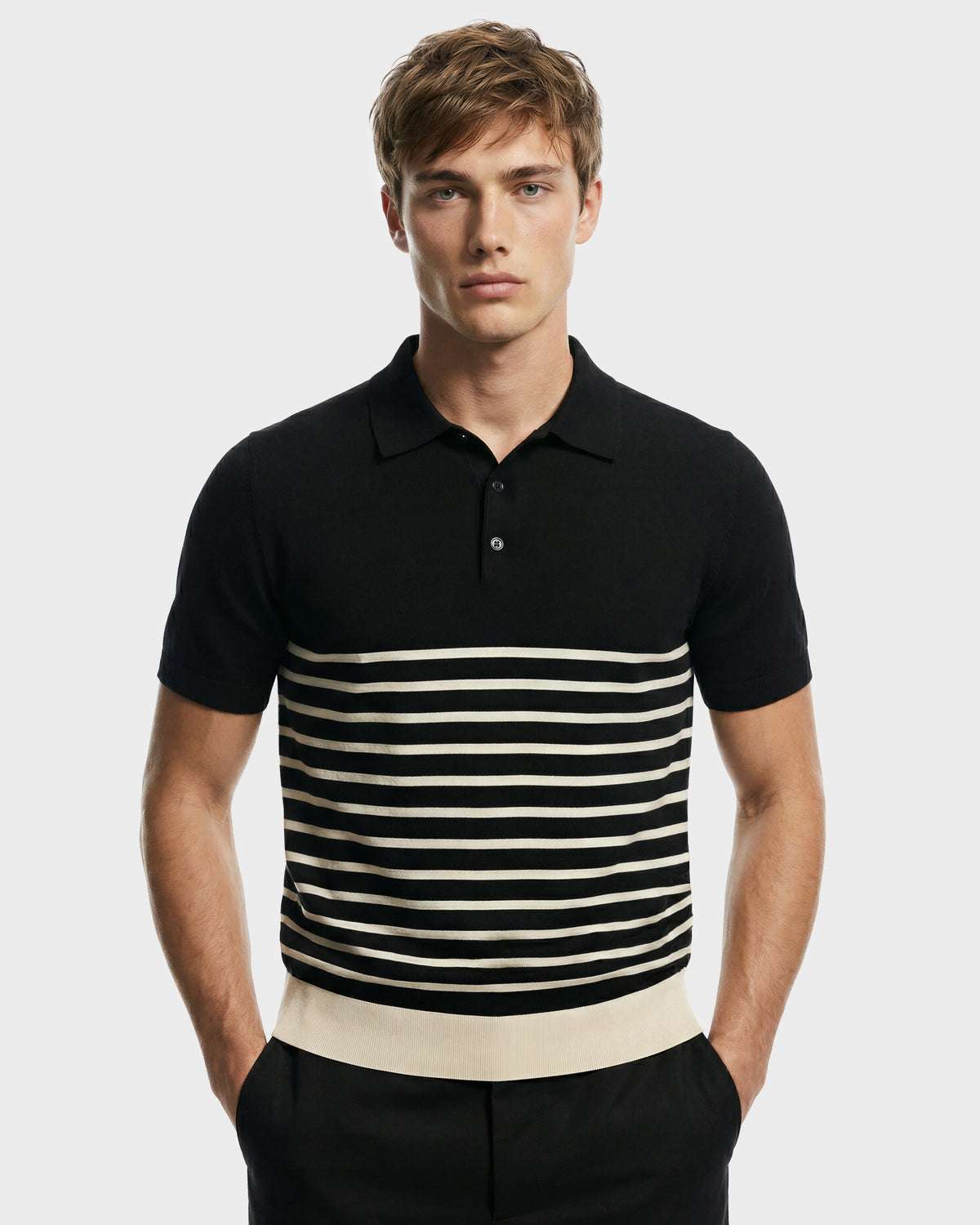 A young man with light brown hair models the KRIS - 6526005 Black men's designer polo by Rudsak, featuring black fabric with white horizontal stripes, standing hands-in-pockets against a plain light gray background.
