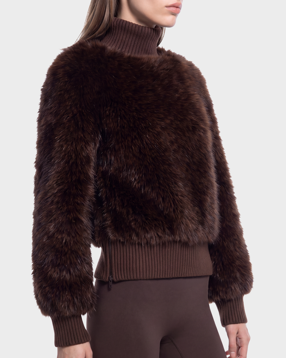 A woman wears the Rudsak LAYLA F - 8525630 Mix Expresso, a dark brown faux fur turtleneck sweater with ribbed sleeves, hem, and neck, styled with matching pants for a cozy après-ski look against a light gray backdrop.