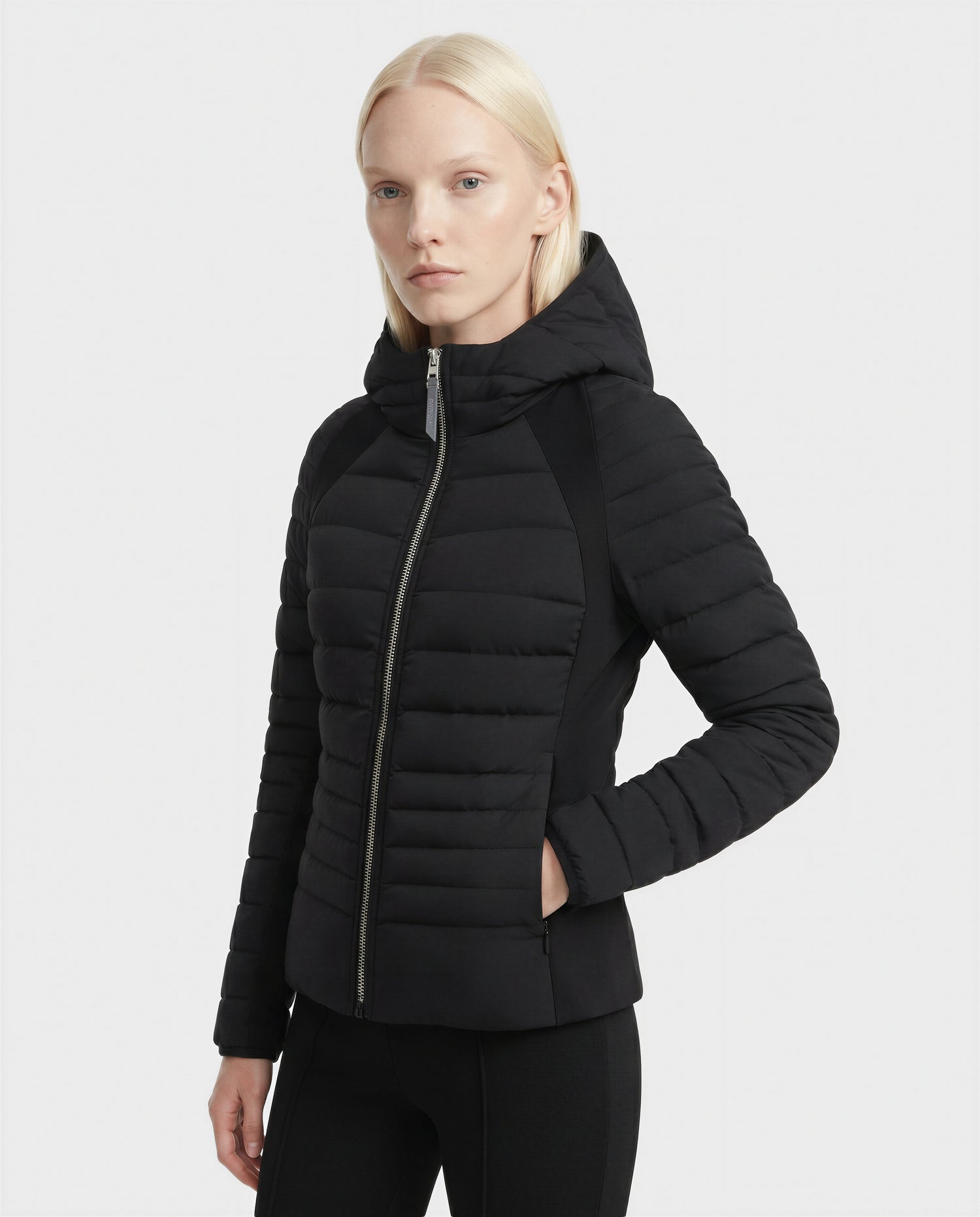 A woman with straight, light blonde hair wears the Rudsak LENA - L124005 Black fitted down jacket with a hood and diagonal front zipper, standing against a white background.