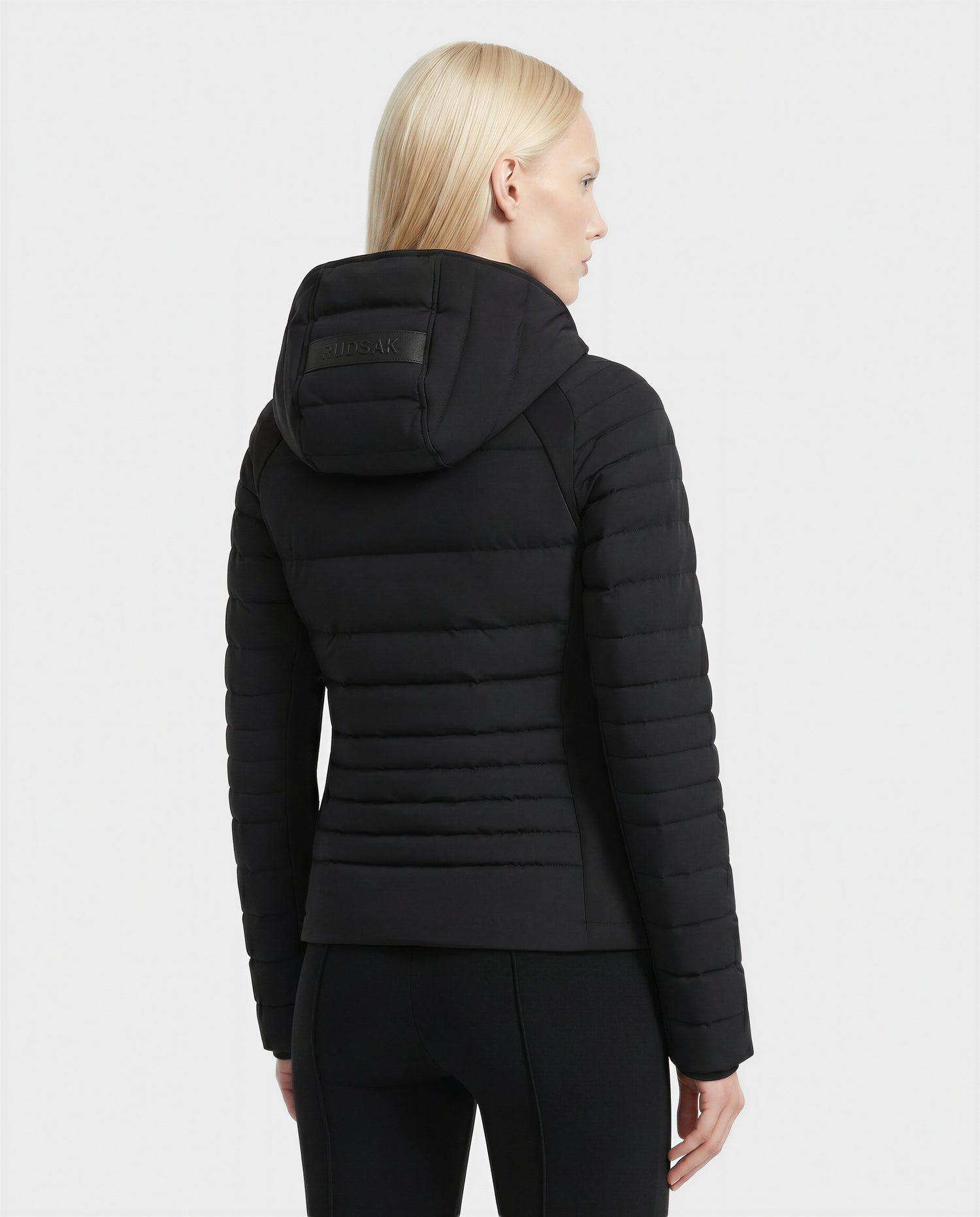 A person with straight, light blonde hair is shown from the back wearing the Rudsak LENA - L124005 Black quilted jacket and black pants against a plain white background.