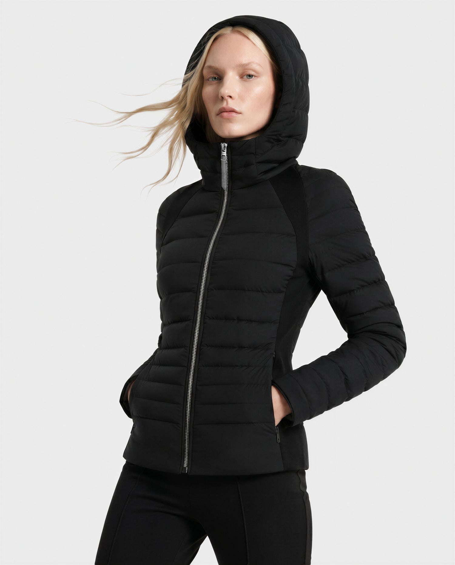 Someone stands with hands in pockets against a plain light background, wearing the Rudsak LENA - L124005 Black fitted quilted jacket featuring a hood and front zipper.