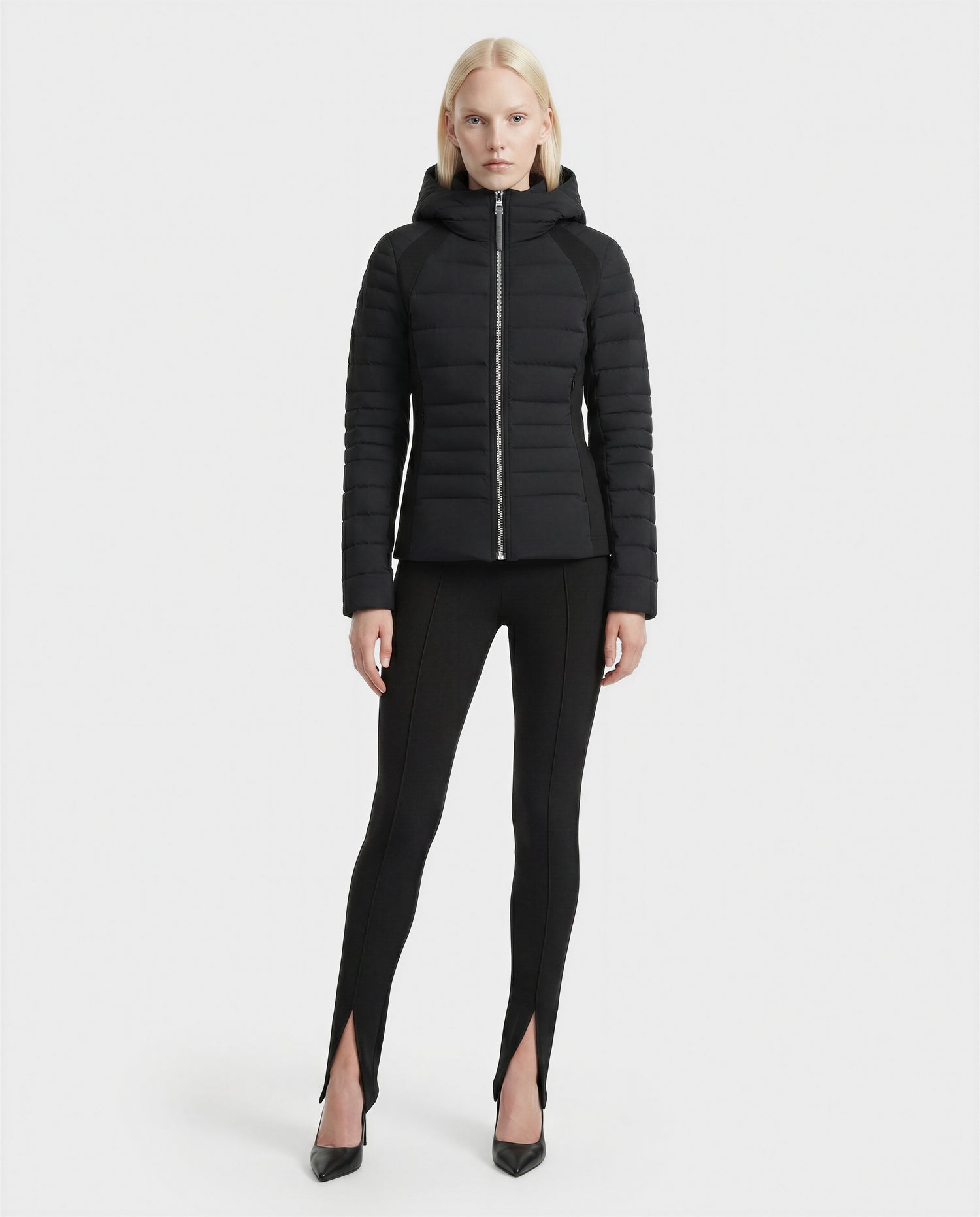 A woman with straight blonde hair wears the Rudsak LENA - L124005 Black fitted quilted jacket, paired with black leggings featuring front ankle slits and black heels, standing against a plain white background.
