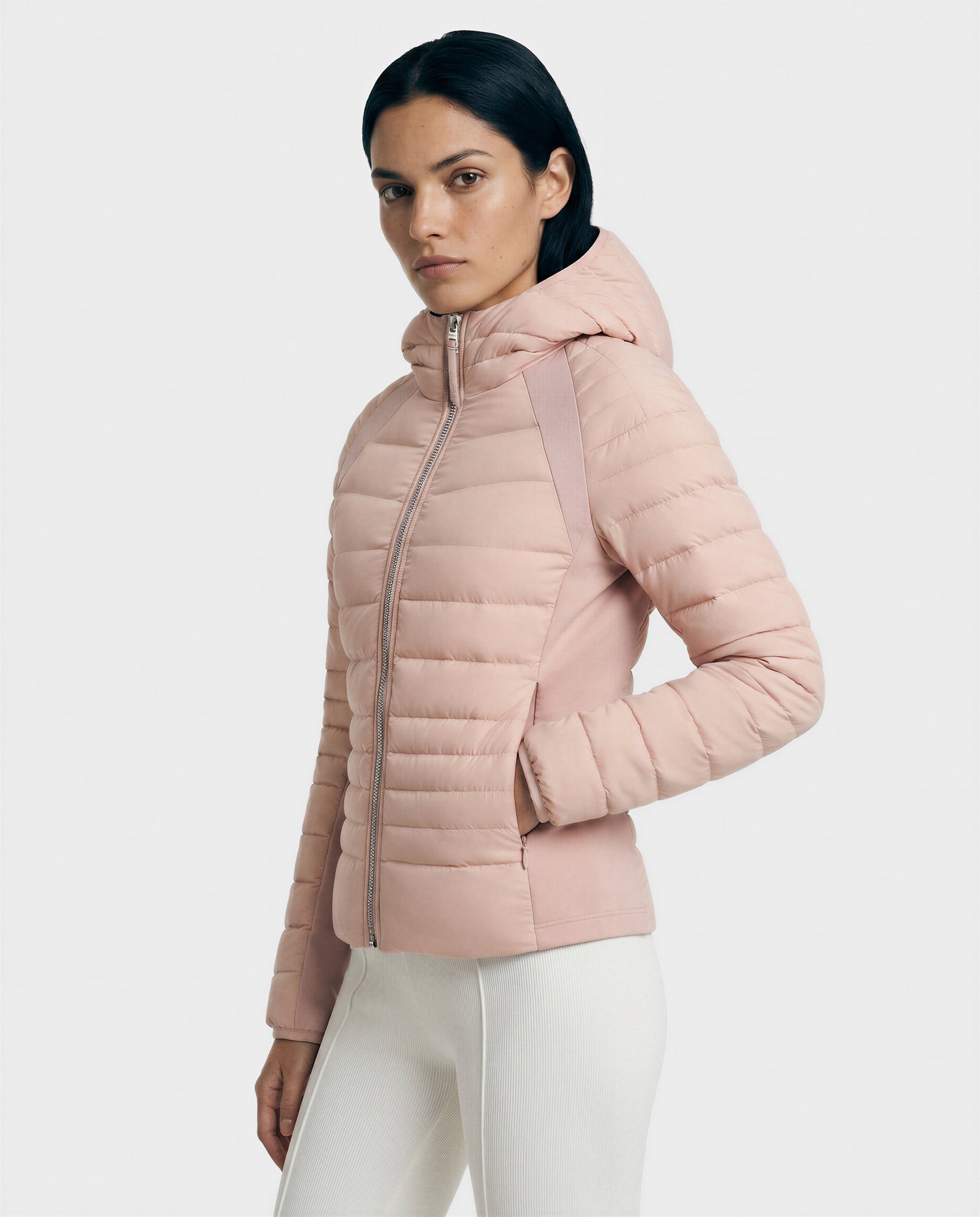 A woman with straight dark hair models the Rudsak LENA - L124005 Soft Pink hooded quilted jacket, paired with white pants, standing against a plain light background and looking at the camera.