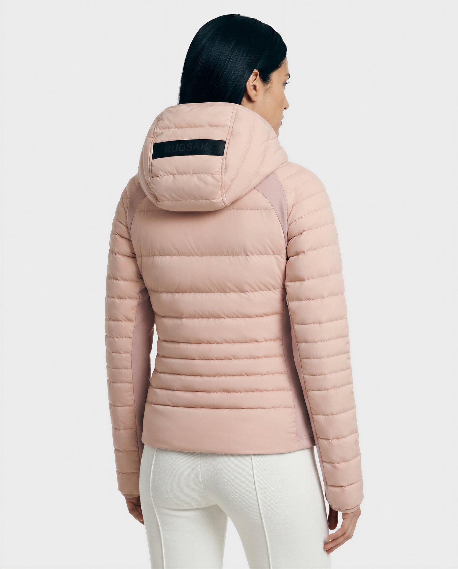 A woman with long dark hair is seen from the back wearing Rudsak's LENA - L124005 quilted light down jacket in soft pink, featuring a hood with a black RUDAK label, and paired with white pants.