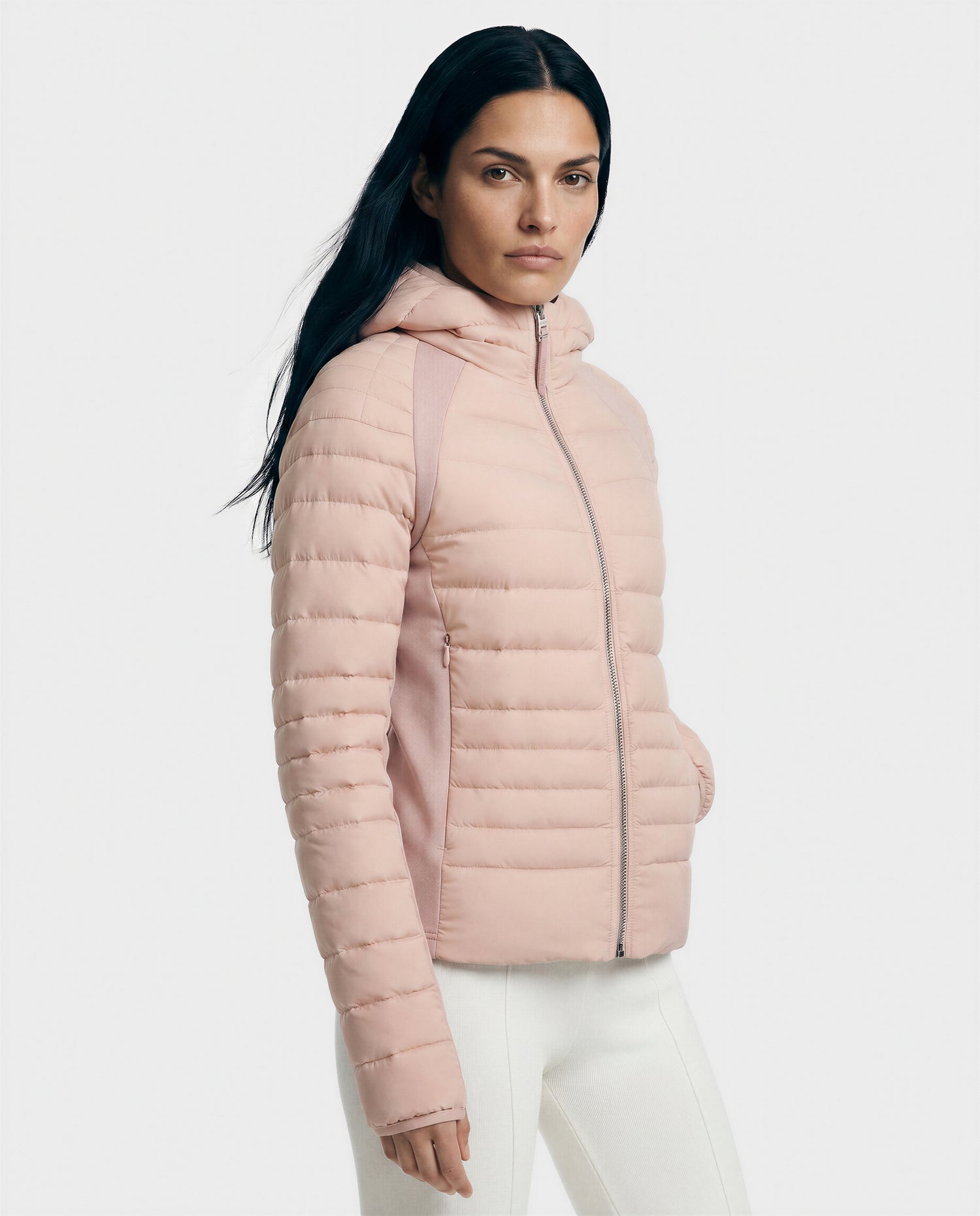 A woman with long dark hair models the Rudsak LENA - L124005 Soft Pink quilted jacket, featuring a hood and front zipper, paired with white pants against a plain light background—a stylish update on the classic down jacket.