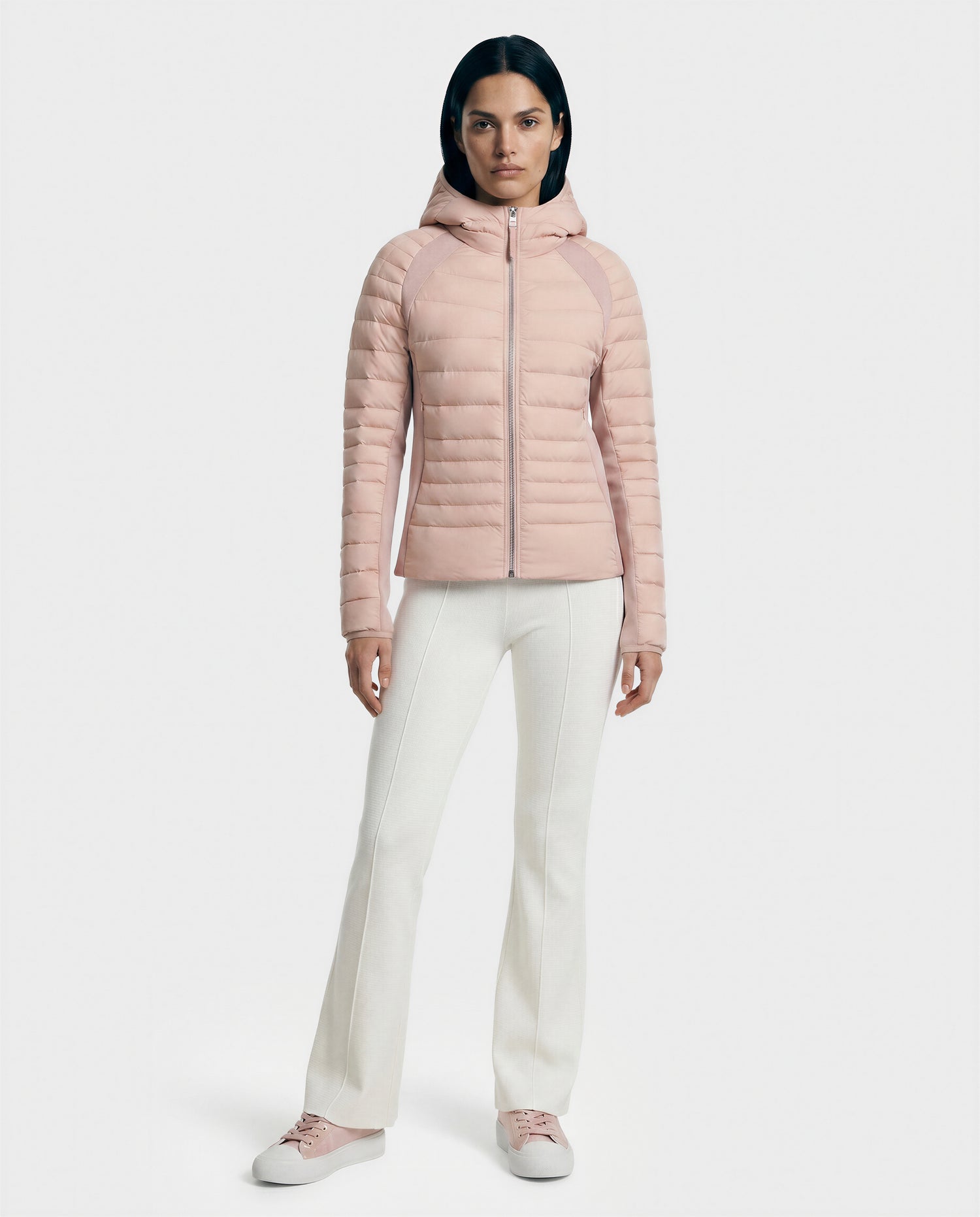 A woman stands facing forward in the Rudsak LENA - L124005 Soft Pink women's light down jacket with subtle neoprene inserts, paired with white pants and light pink sneakers, against a plain white background.