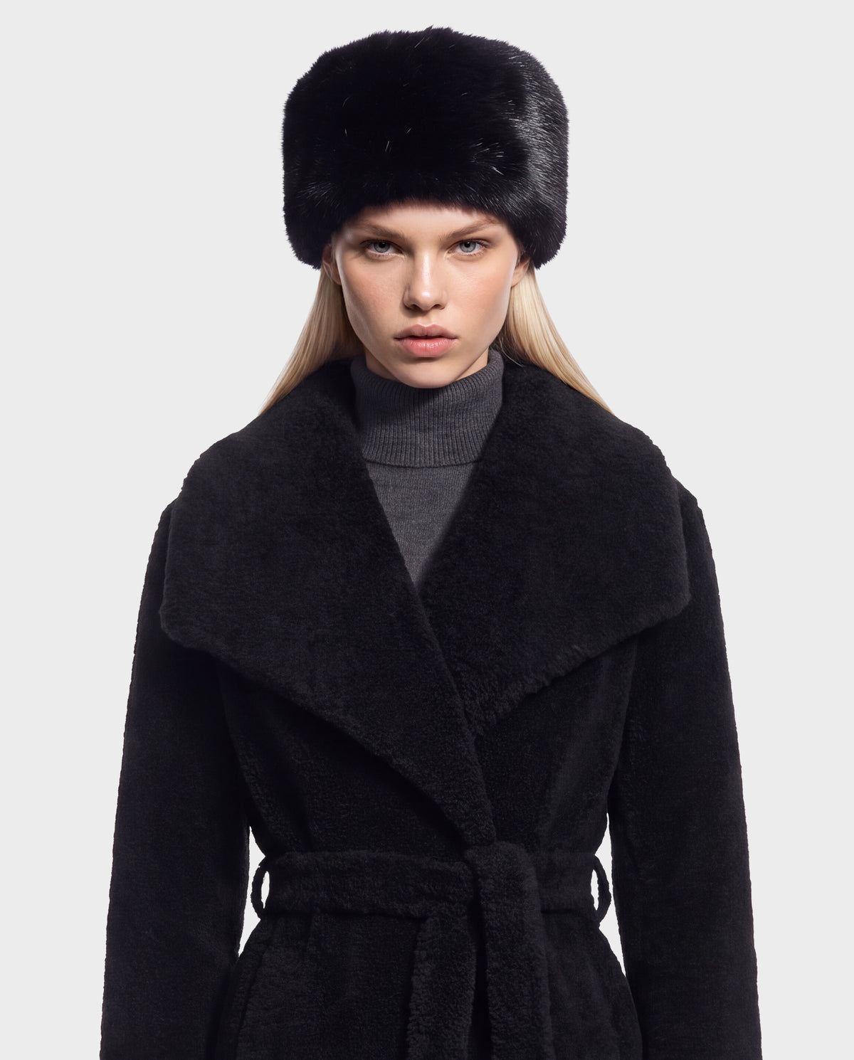 A person wearing a black fur hat and a LORETTA - 8426503 Black belted coat with a large collar over a gray turtleneck sweater.
