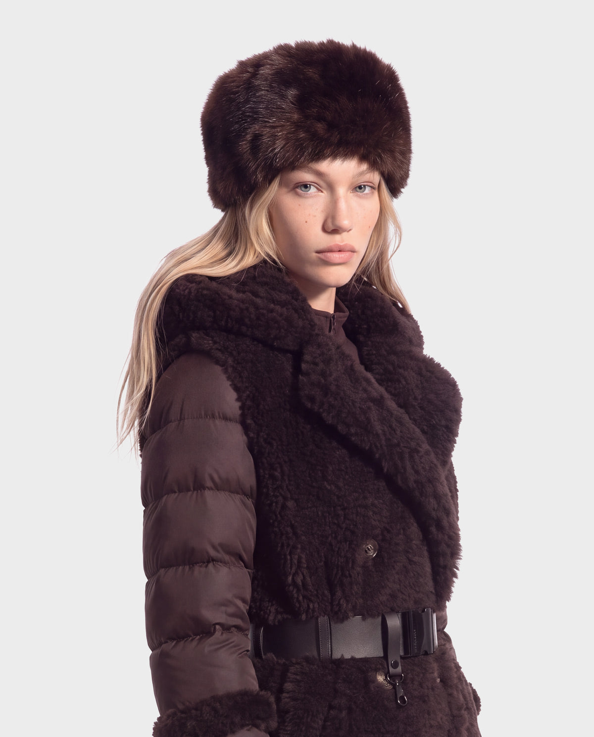 A person wearing a LORETTA - 8426503 Mix Espresso jacket with a dark brown, fur-trimmed hat, a quilted puffer design with a faux-fur front panel, and a black belt.