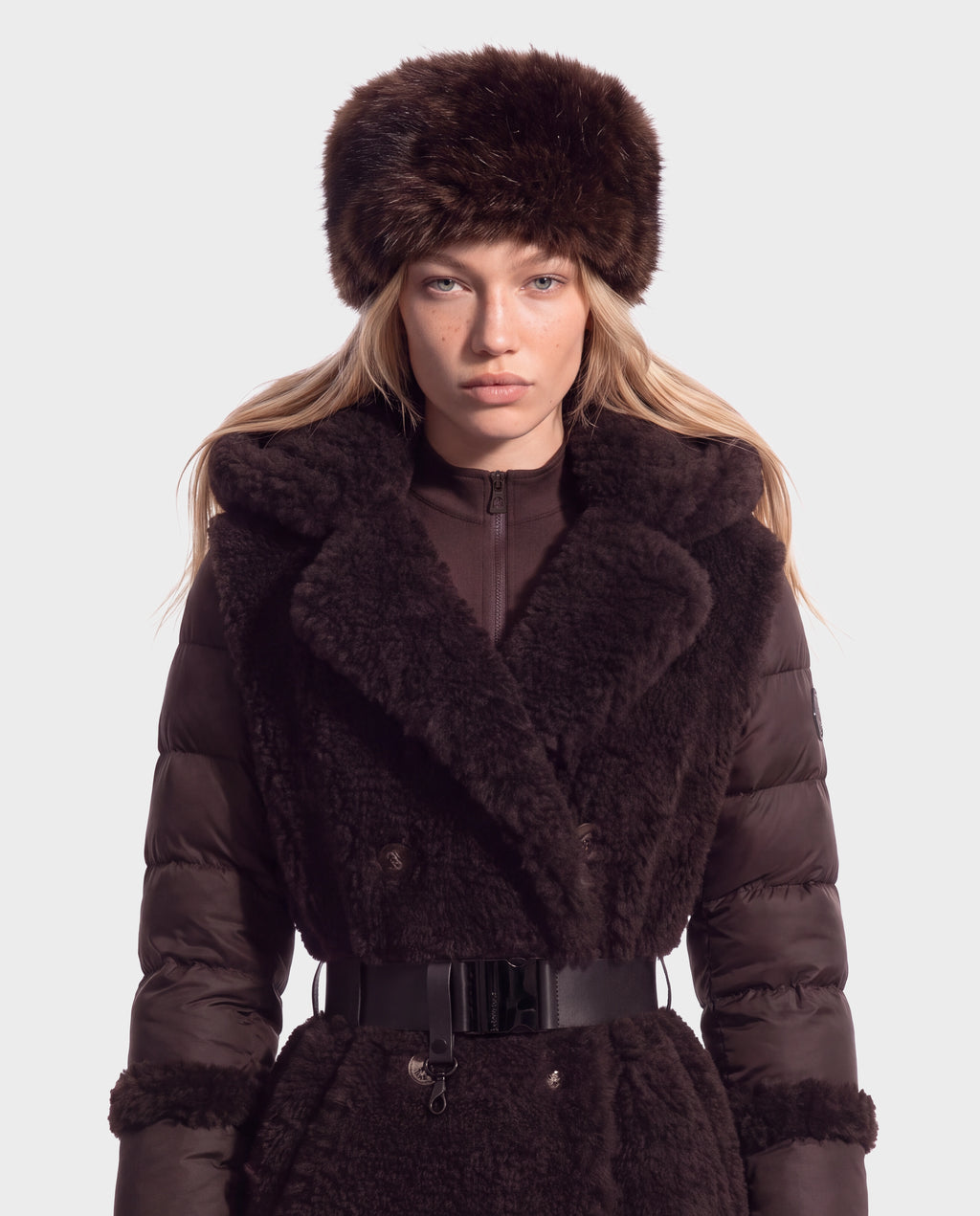 A person wearing a LORETTA - 8426503 Mix Espresso dark brown fur hat and a matching puffer coat with fur trim and a belted waist.
