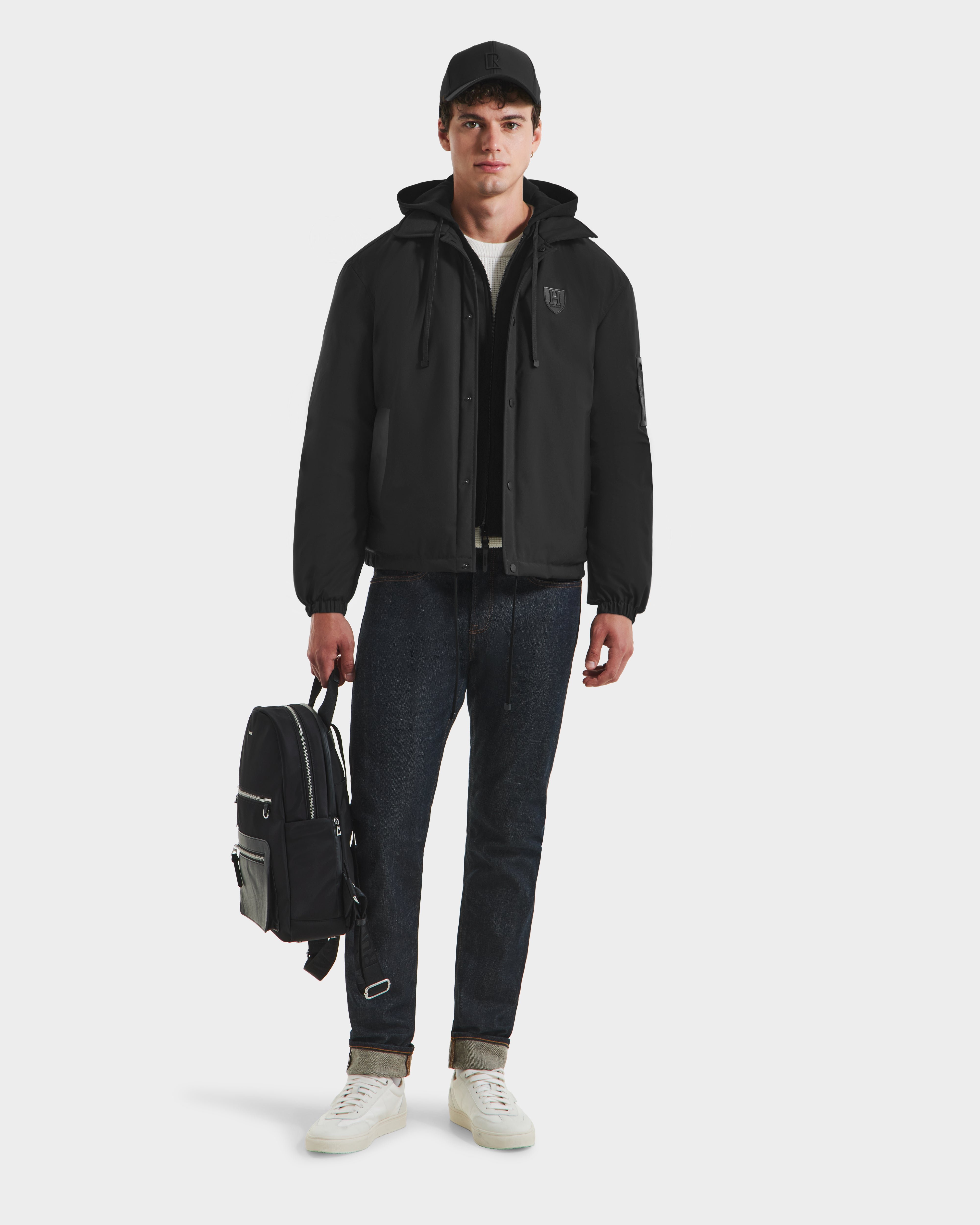 A young man wearing the Rudsak LUCA HV - H125505 Black outerwear, black cap, dark jeans, and white sneakers stands facing forward, holding a black backpack in his left hand against a plain white background.