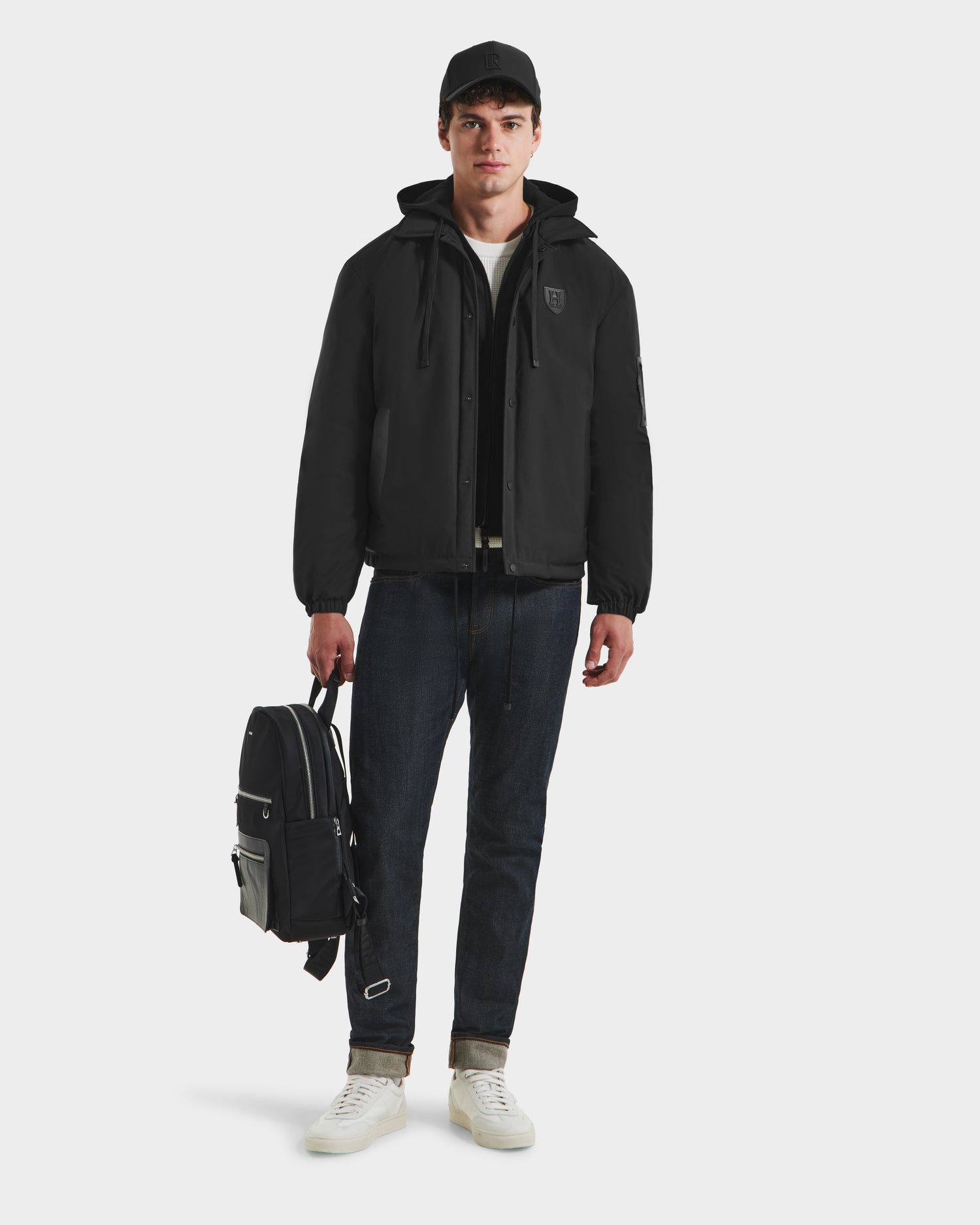 A young man wearing the Rudsak LUCA HV - H125505 Black outerwear, black cap, dark jeans, and white sneakers stands facing forward, holding a black backpack in his left hand against a plain white background.