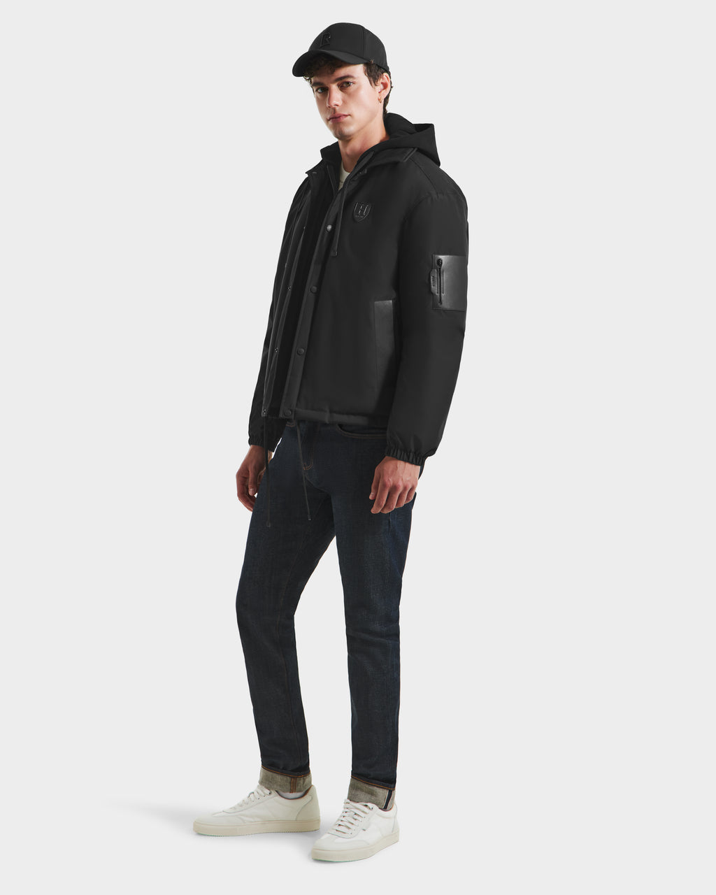 A young man in a black cap, Rudsak LUCA HV - H125505 Black limited-edition outerwear, dark jeans, and white sneakers stands slightly turned against a plain white background.