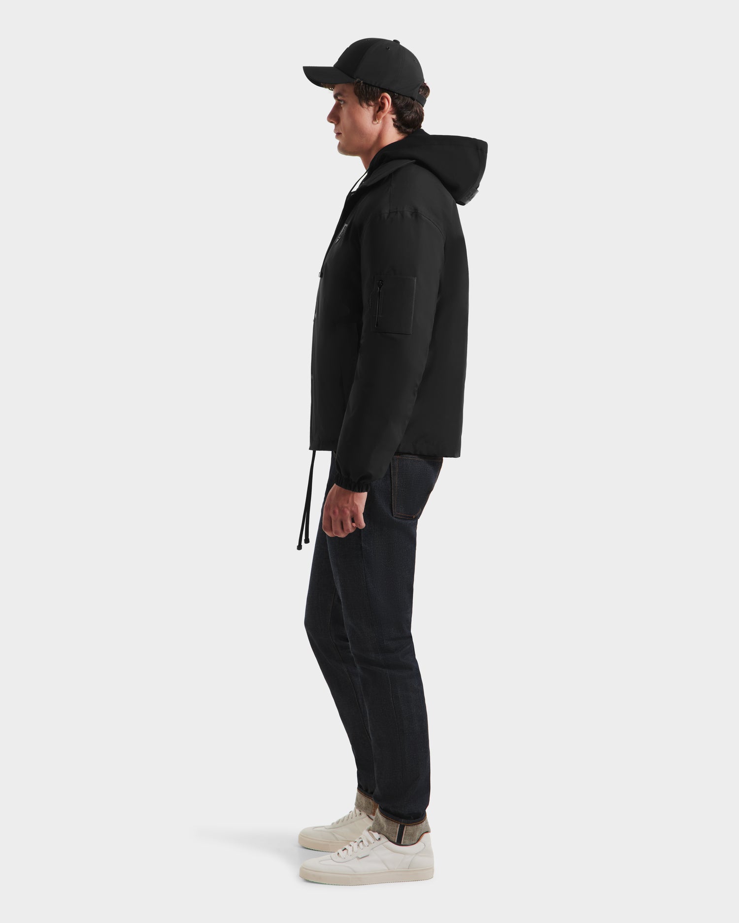 A man in a Rudsak LUCA HV - H125505 Black limited-edition hooded jacket, black cap, dark jeans, and white sneakers stands in profile facing left against a plain white background.