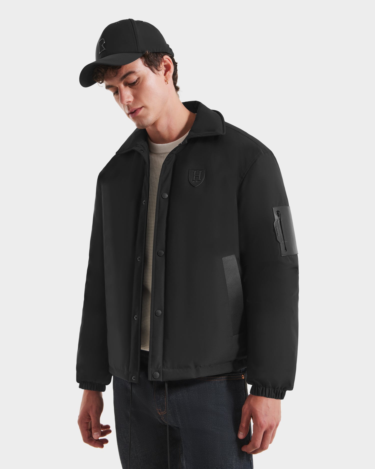 A young man wears the Rudsak LUCA HV - H125505 Black jacket, a black button-up limited-edition piece, paired with a black baseball cap, beige ribbed sweater, and dark jeans while standing against a white background.