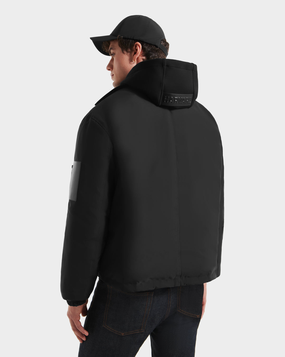 A person wearing the Rudsak LUCA HV - H125505 Black puffer jacket from the Harvard x RUDSAK limited-edition collection, with a high collar and white sleeve patch, is shown from behind in a black cap and dark jeans against a plain backdrop.