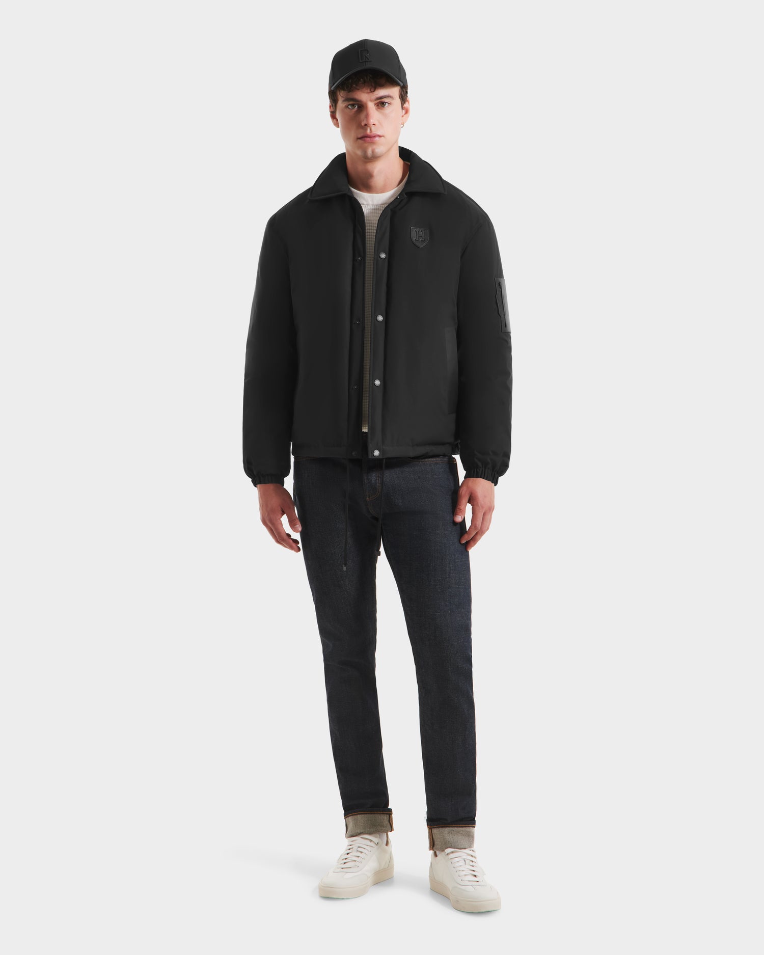 A young man stands facing forward, wearing the Rudsak LUCA HV - H125505 Black jacket, a black baseball cap, cuffed dark jeans, white t-shirt, and white sneakers against a plain light background.