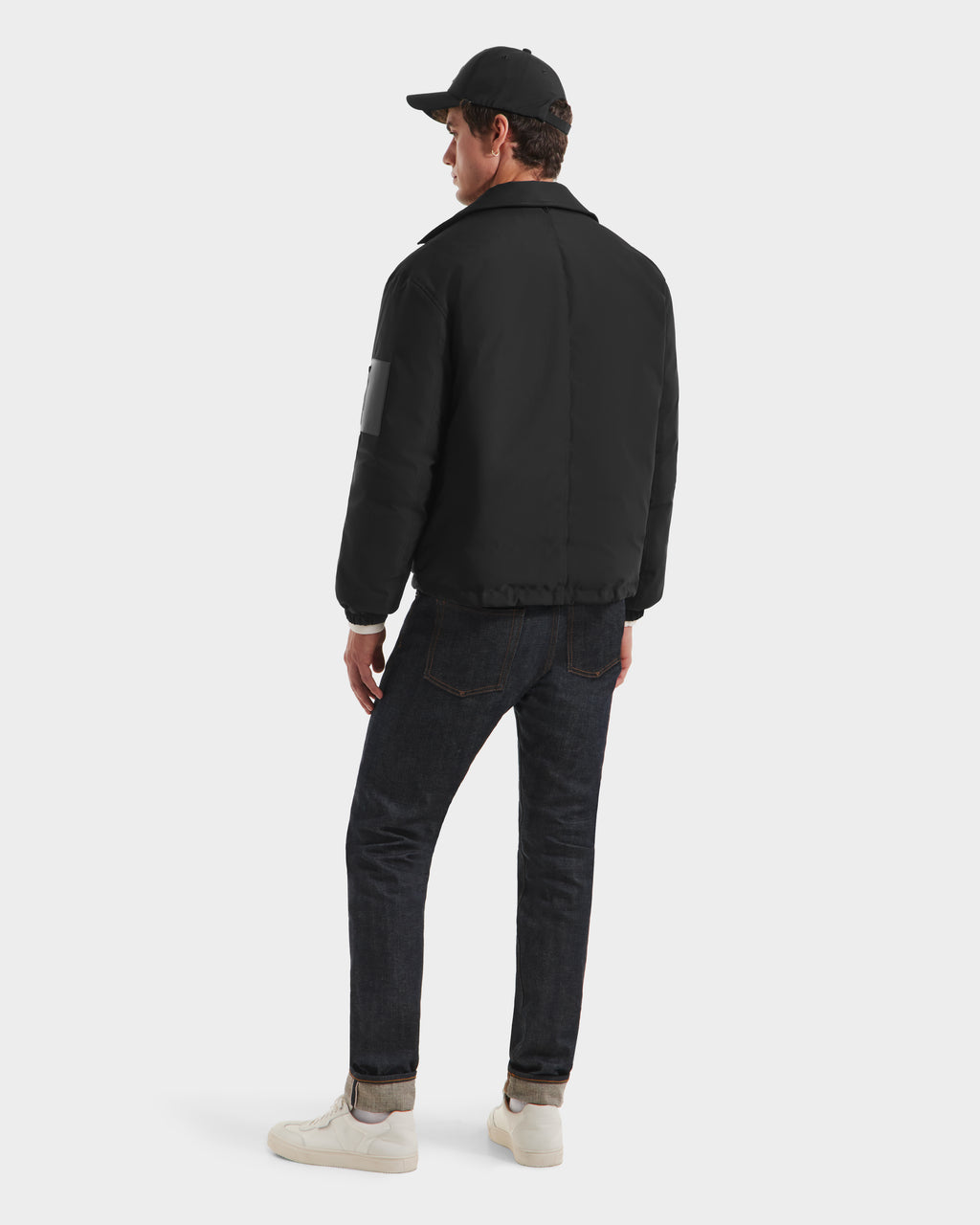 A man wearing the Rudsak LUCA HV - H125505 Black jacket, dark jeans, white sneakers, and a black baseball cap stands facing away from the camera against a plain white background.