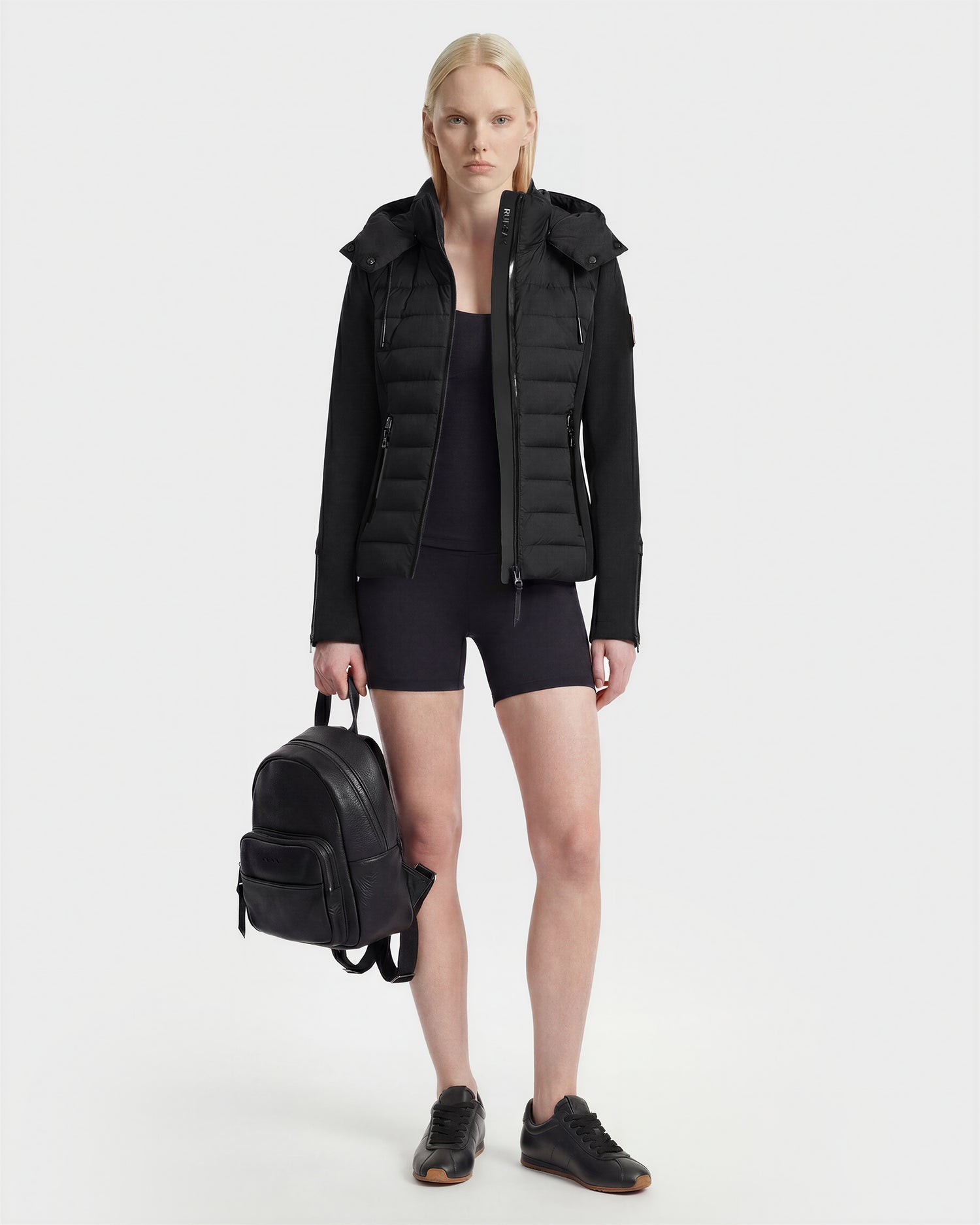 A woman with straight blonde hair wears the Rudsak MABS - 8125055 Black puffer jacket, paired with black shorts, black sneakers, and stands beside a black backpack against a plain white background.
