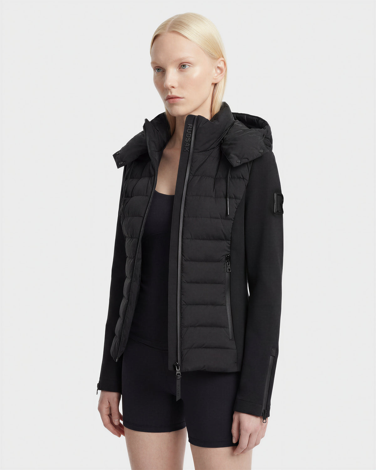 A woman with straight blonde hair wears the Rudsak MABS - 8125055 Black women's puffer jacket with a hood over a black top and shorts, standing against a plain light background.