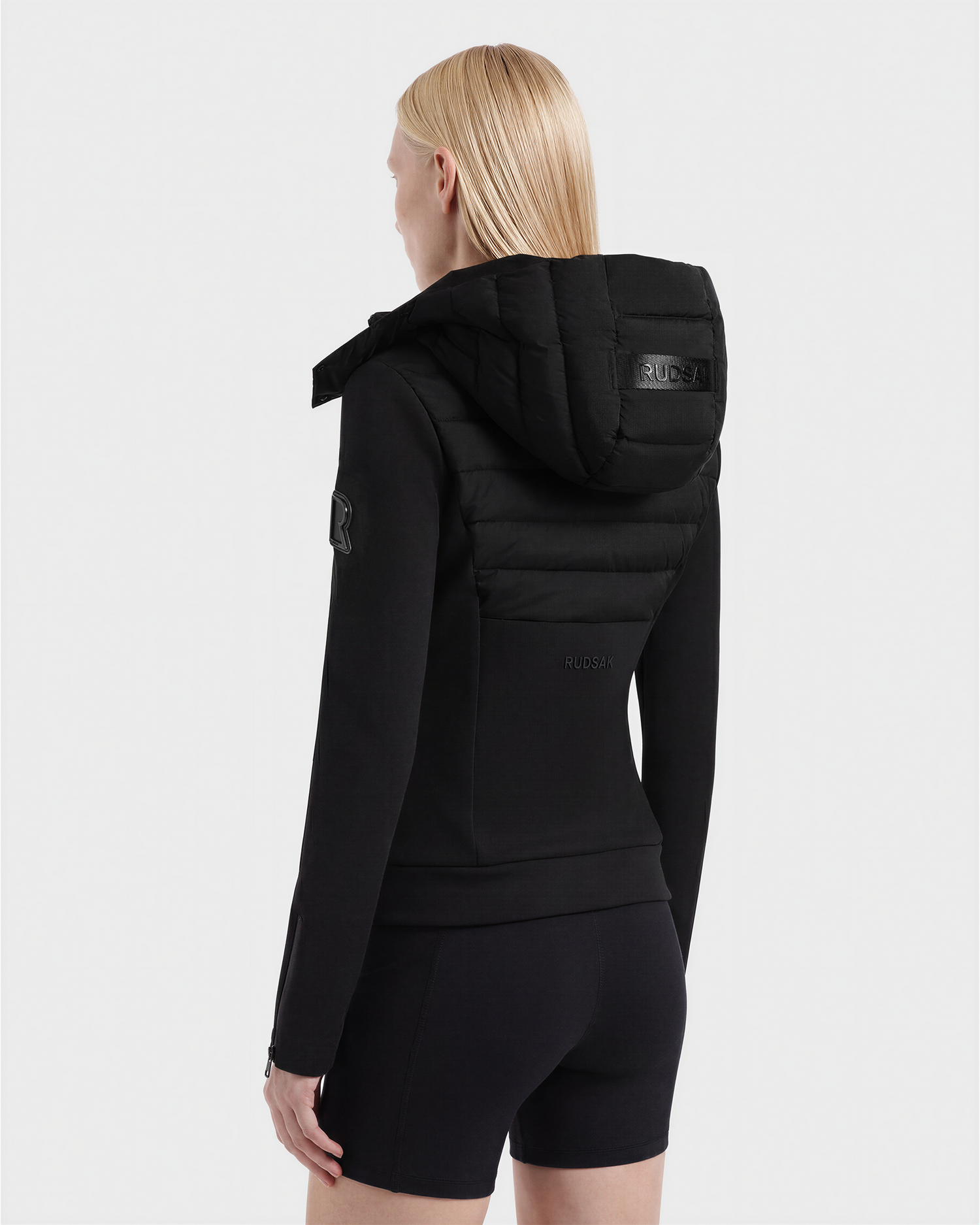 A person with straight blonde hair wears the Rudsak MABS - 8125055 Black women's puffer jacket and black shorts, standing with their back to the camera against a plain light background.