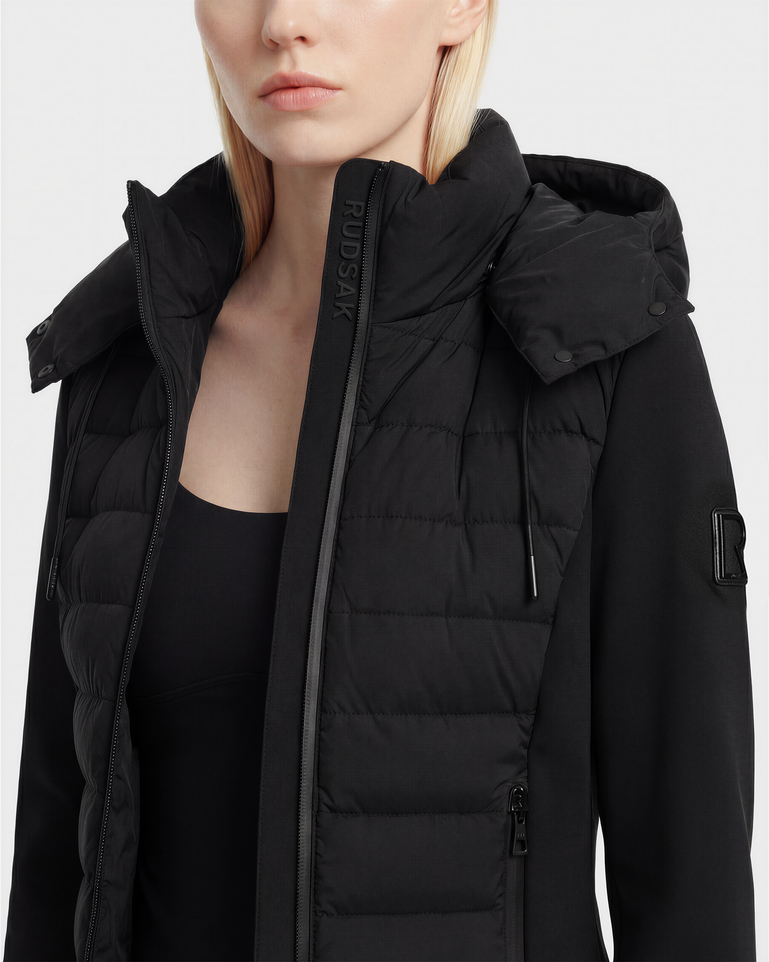 A woman with straight light blonde hair and a neutral expression wears the Rudsak MABS - 8125055 Black women’s puffer jacket with a high collar and hood, made from RDS-certified down, standing against a plain white background.