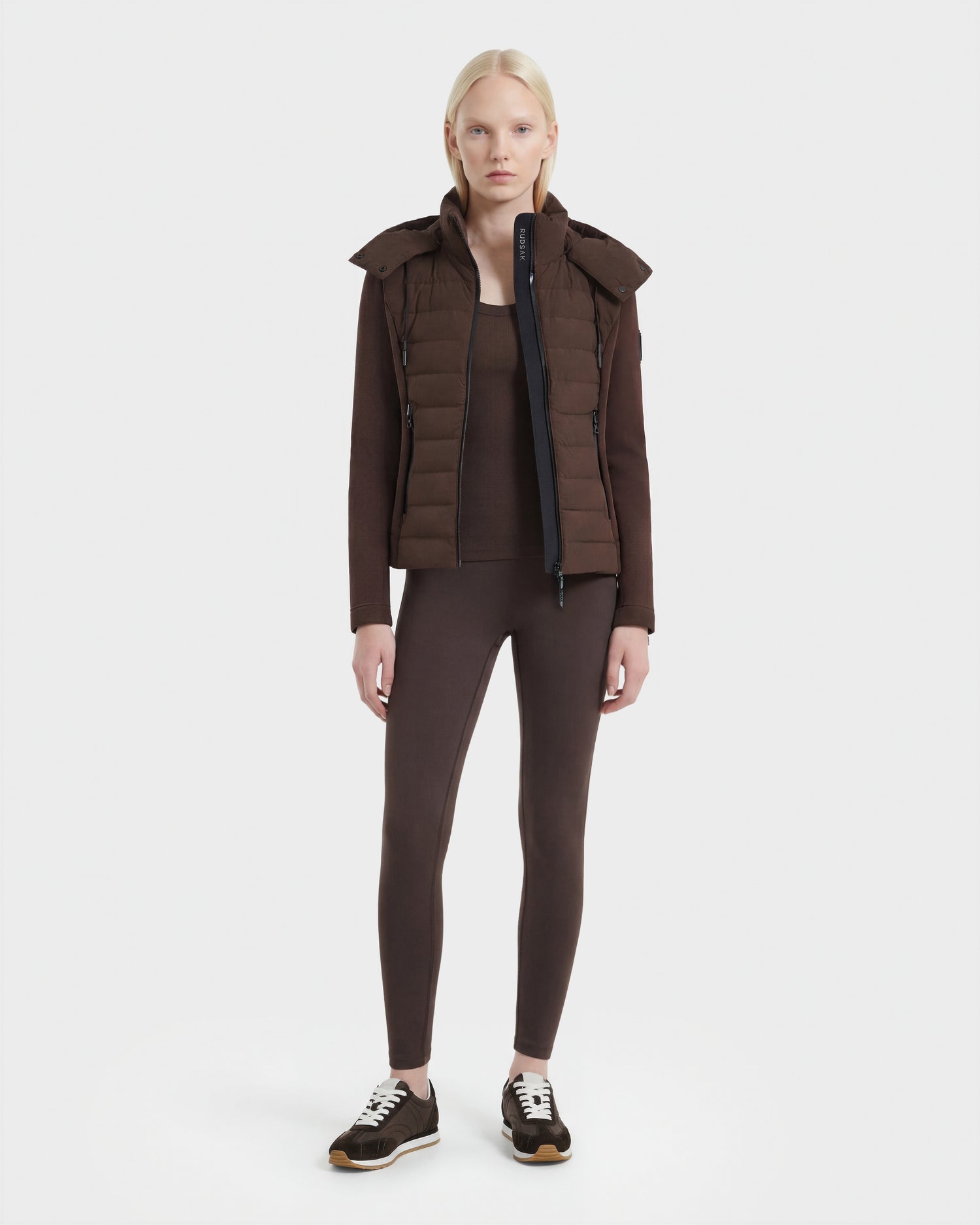 A woman with long blonde hair stands facing forward, wearing the Rudsak MABS - 8124825 Espresso neoprene short jacket over a matching top and leggings, paired with black sneakers, against a plain white background.
