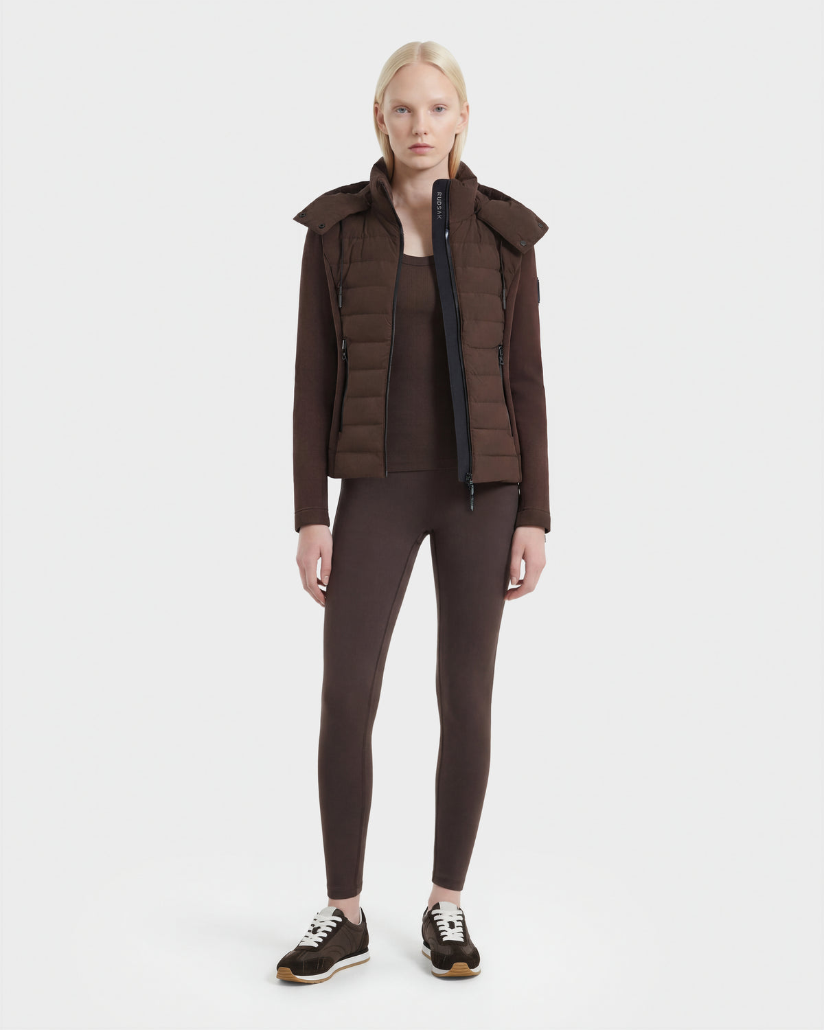 A woman with long blonde hair stands facing forward, wearing the Rudsak MABS - 8124825 Espresso neoprene short jacket over a matching top and leggings, paired with black sneakers, against a plain white background.