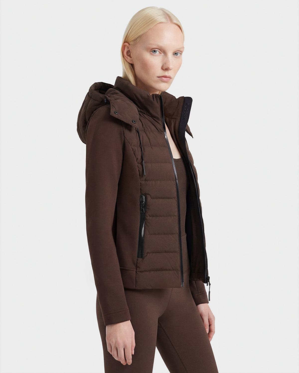 A woman with straight, light blonde hair models the Rudsak MABS - 8124825 Espresso fitted women's puffer jacket featuring a black zipper and hood, paired with matching brown leggings against a plain white background.