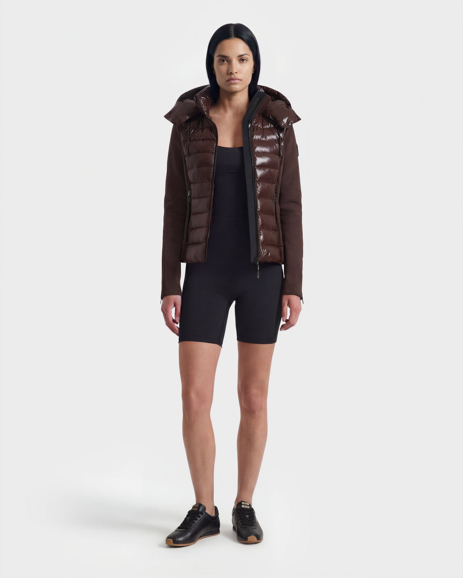 A woman faces forward against a white background, wearing the Rudsak MABS C - 8124590 Espresso shiny dark brown puffer jacket over a black fitted romper and black sneakers.