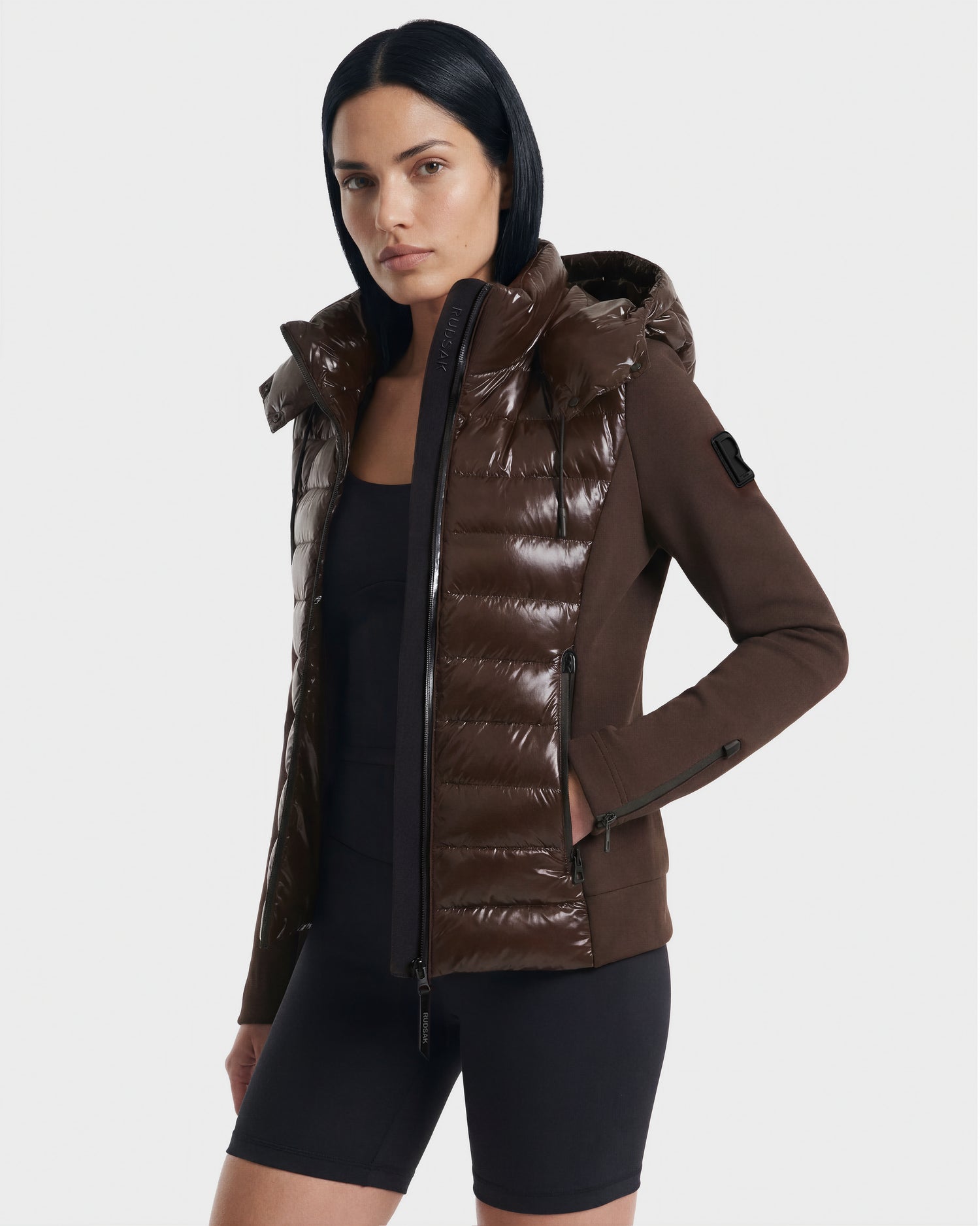 A woman with straight black hair models the Rudsak MABS C - 8124590 Espresso women's activewear jacket over a black top and bike shorts, standing against a plain light background.
