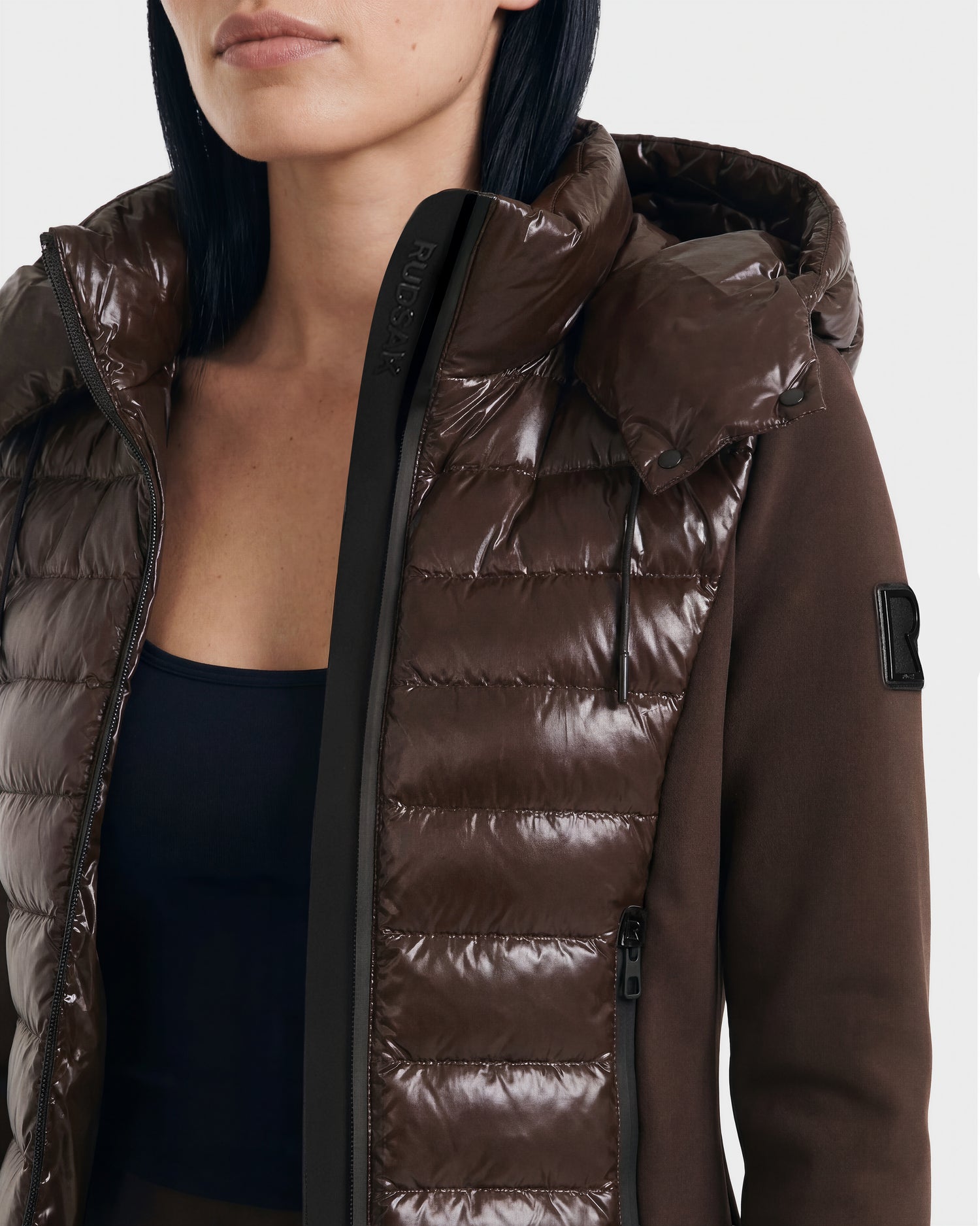A woman with straight black hair wears the Rudsak MABS C - 8124590 Espresso, a shiny brown puffer jacket with a hood. The lightweight jacket is partially unzipped to reveal a black top and has a visible zipper and patch on the sleeve.