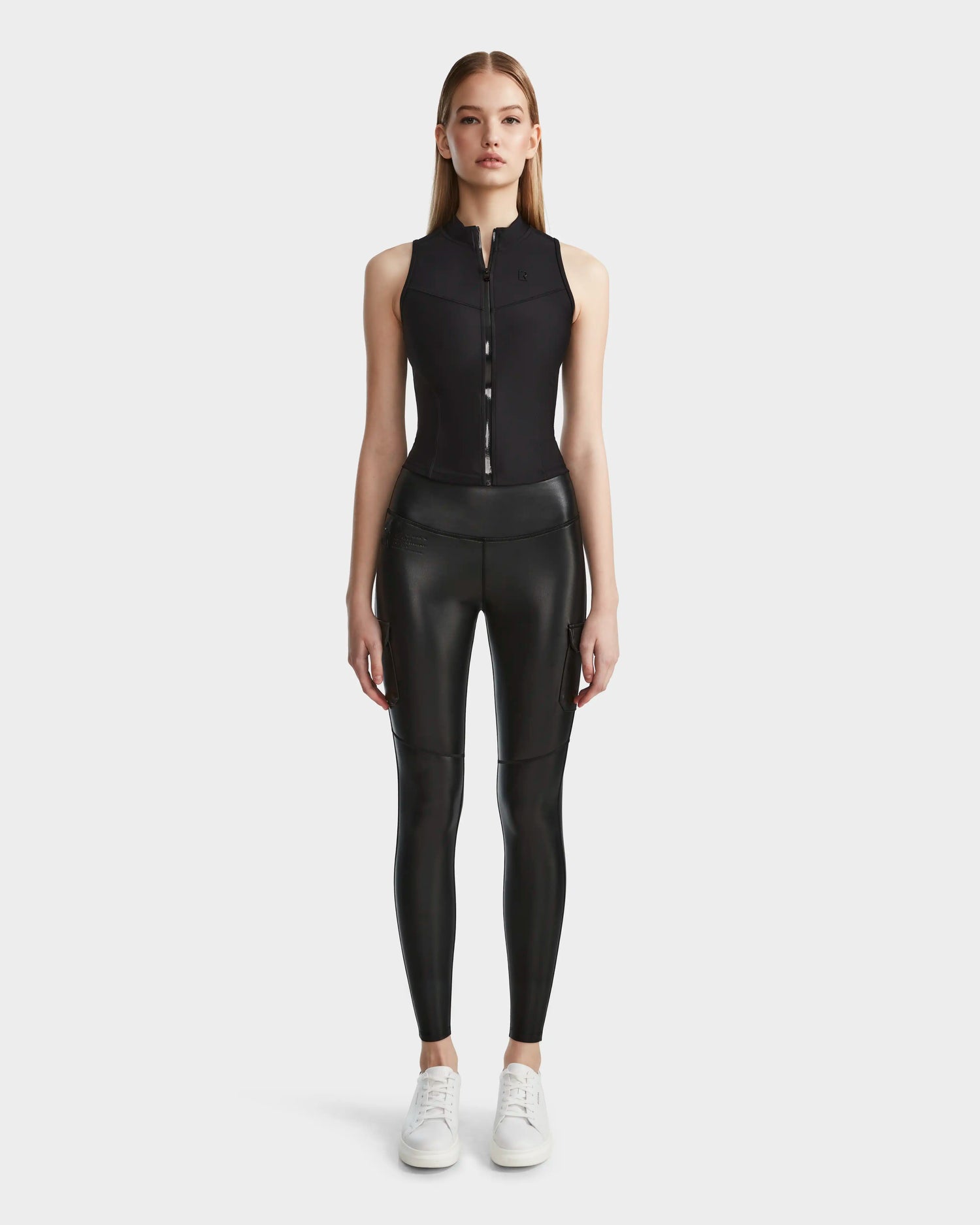 A woman faces forward, wearing the Rudsak SIEN L - 8524079 Black sleeveless zip-up top and matching black high-waisted vegan leather leggings with white sneakers against a plain light background.