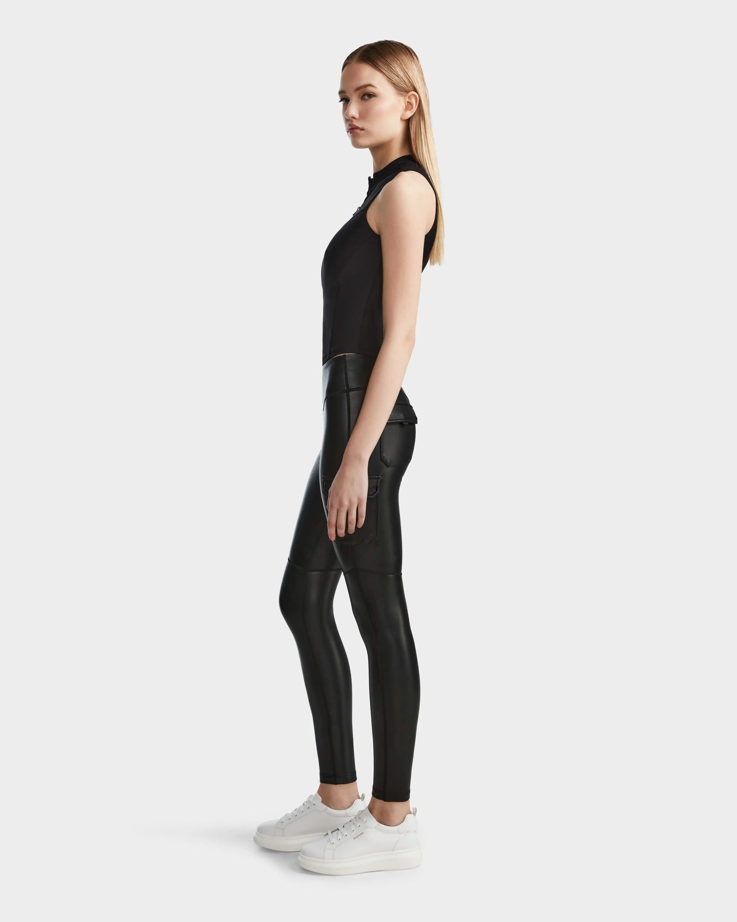 A woman with straight blond hair and a neutral expression stands sideways against a plain light background, wearing Rudsak's SIEN L - 8524079 Black vegan leather leggings and a sleeveless black top, paired with white platform sneakers.
