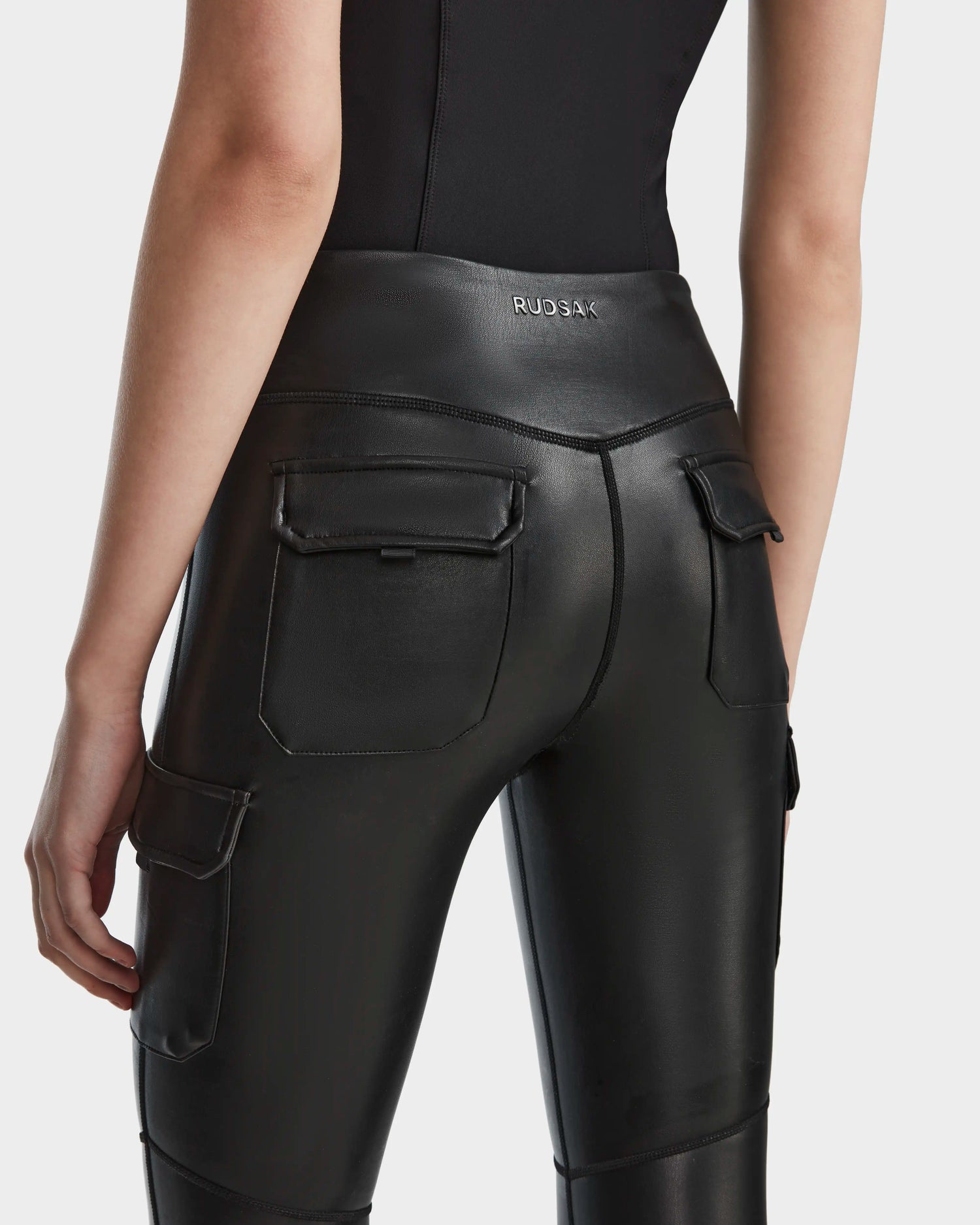 A person shown from the waist down and viewed from behind wearing Rudsak's SIEN L - 8524079 Black fitted vegan leather leggings featuring back and side pockets, cargo pocket detailing, and a visible RUDSAK logo on the waistband.