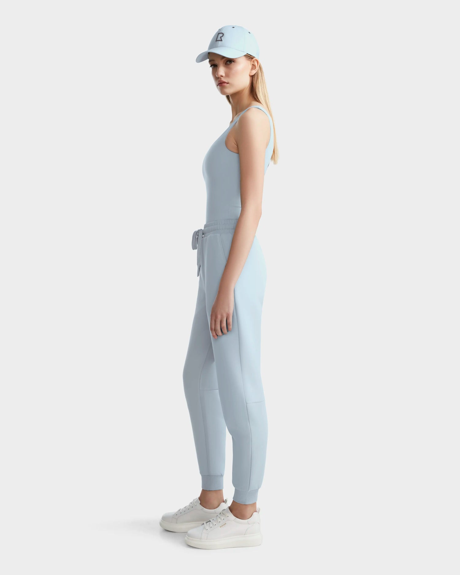 A woman models the Rudsak BRIDGET - 8524033 Sky Blue tank bodysuit with jogger pants, a matching sky blue cap, and white sneakers against a white background for a sporty-chic look.