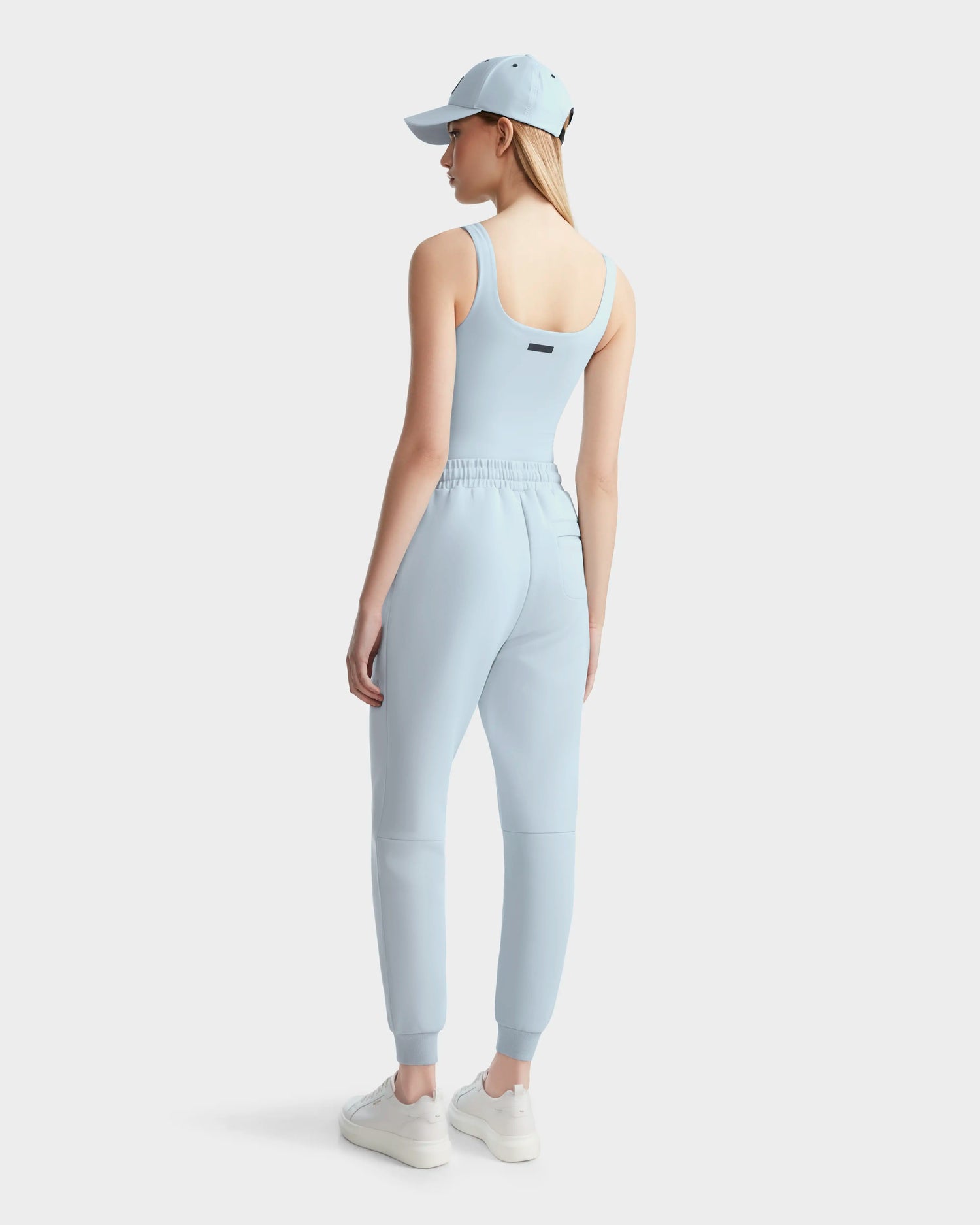 A woman wears the Rudsak BRIDGET - 8524033 Sky Blue stretch tank bodysuit, jogger pants, a baseball cap, and white sneakers, standing against a plain white background and looking to the side.