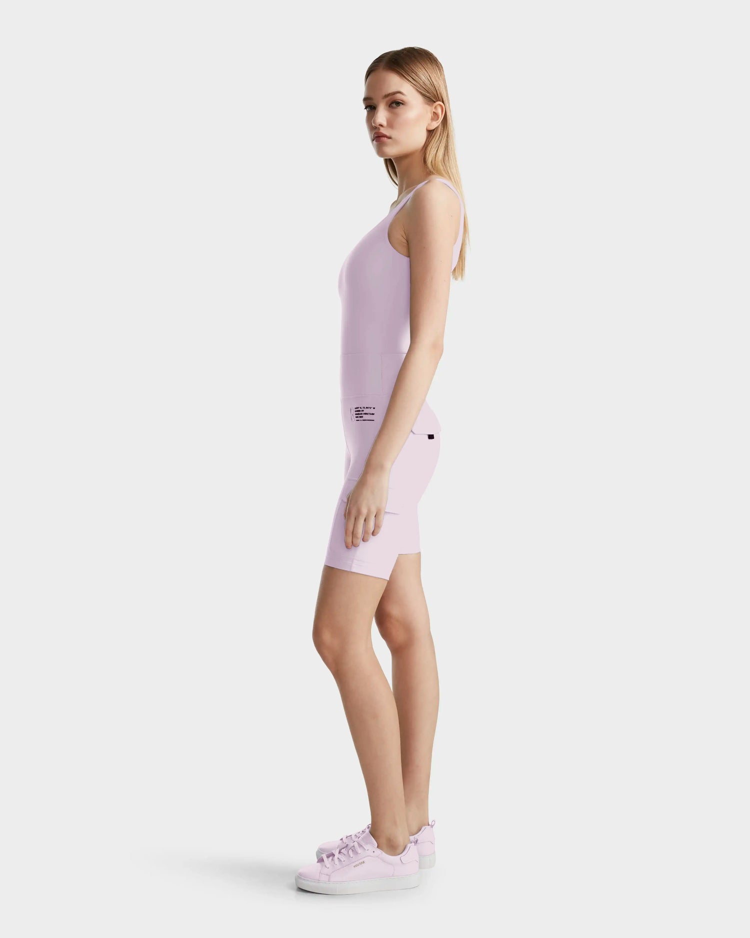 A woman stands sideways wearing the Rudsak ZORA - 8524066 Lilac Foam sleeveless fitted romper, crafted from high-performance stretch fabric, paired with matching pink sneakers against a plain white background.