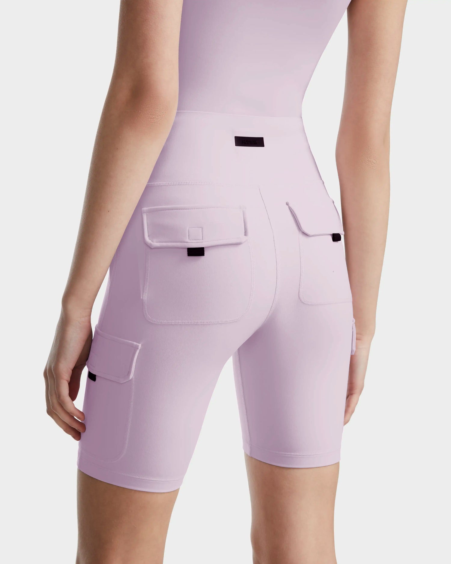 A woman wears Rudsak's ZORA - 8524066 Lilac Foam high-waisted biker shorts with multiple cargo pockets, shown from the back against a white background.