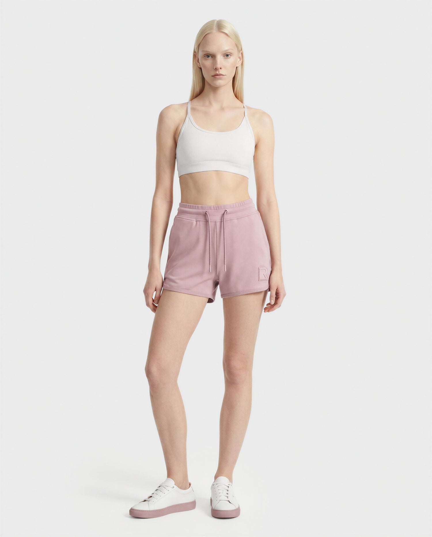 A woman faces forward against a white background, wearing a white sports bra, Rudsak SAMMY - L523014B soft pink drawstring shorts, and white sneakers with pink soles.