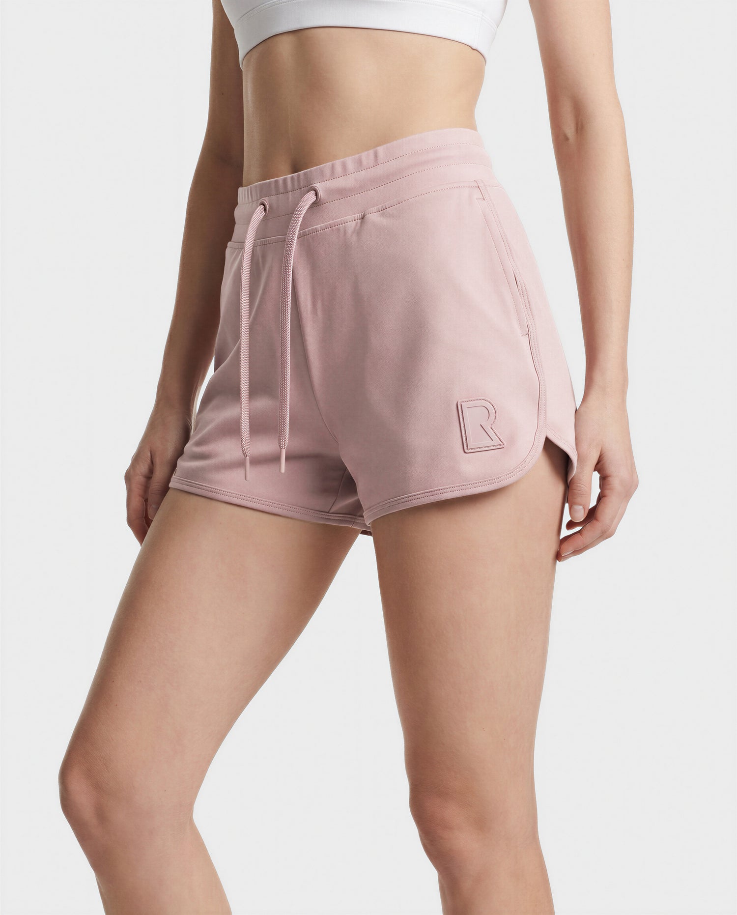 A person wears Rudsak's SAMMY - L523014B Bottom in Soft Pink, featuring a drawstring and raised “R” logo on the left leg, paired with a white sports bra, against a plain white background.