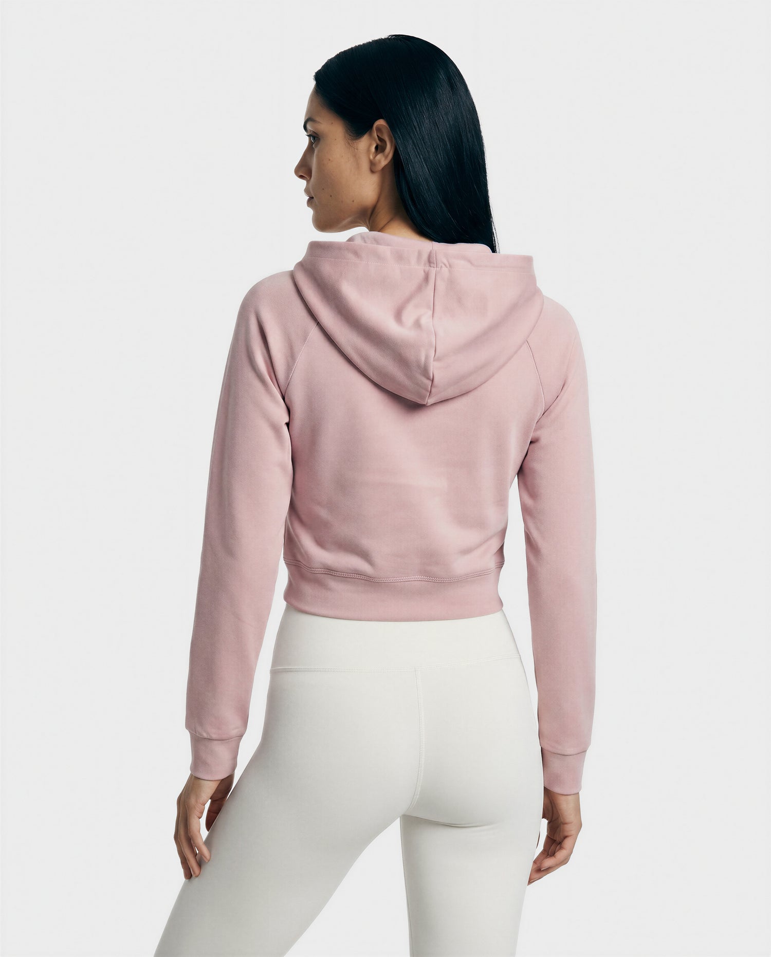 A woman with long dark hair, seen from behind, wears the Rudsak SAMMY - L523014T Top in Soft Pink and high-waisted white leggings, standing against a plain light gray background.