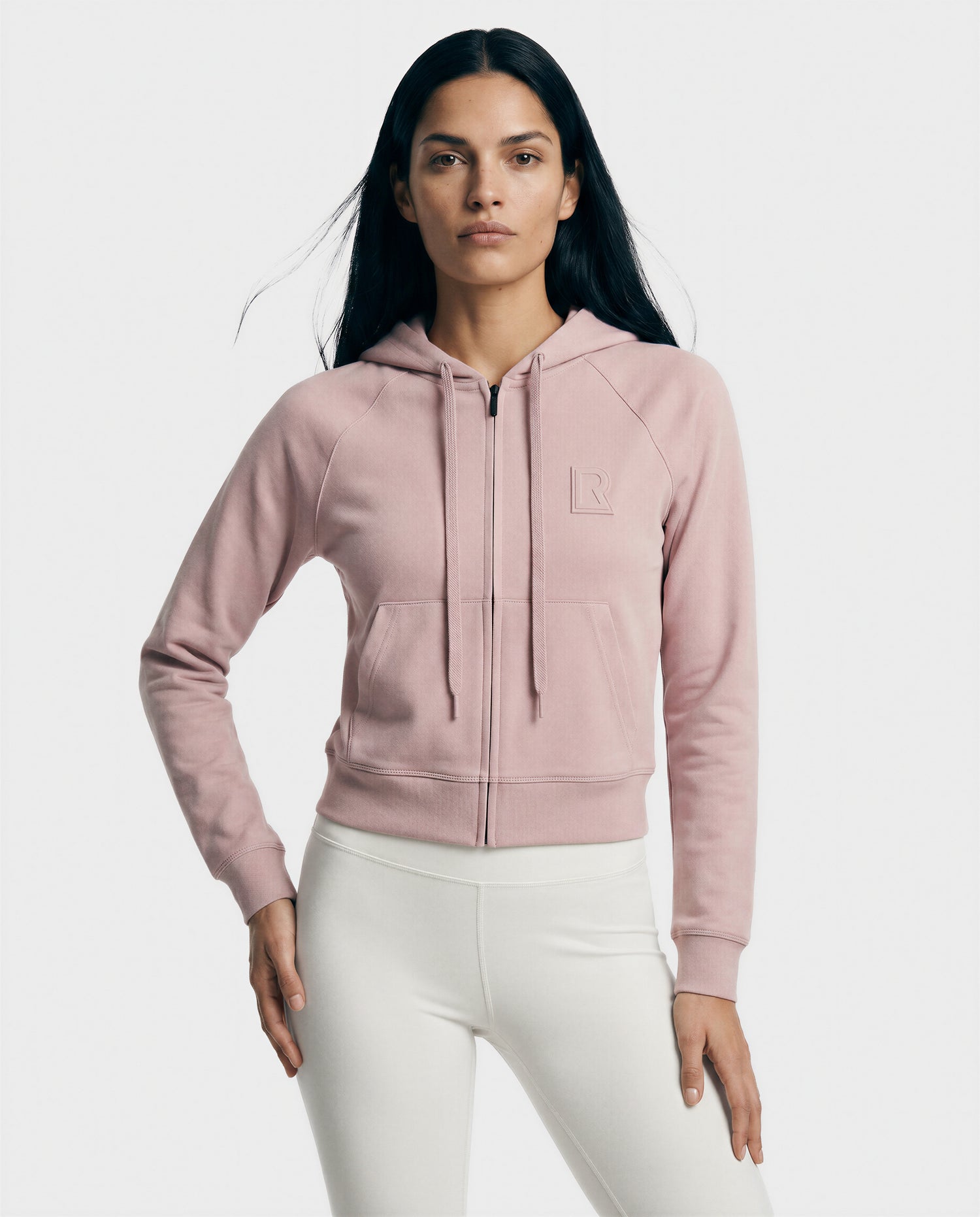 A woman with long dark hair models the Rudsak SAMMY - L523014T top in soft pink, featuring a zip-up front and small chest logo, paired with white fitted pants against a plain light background.