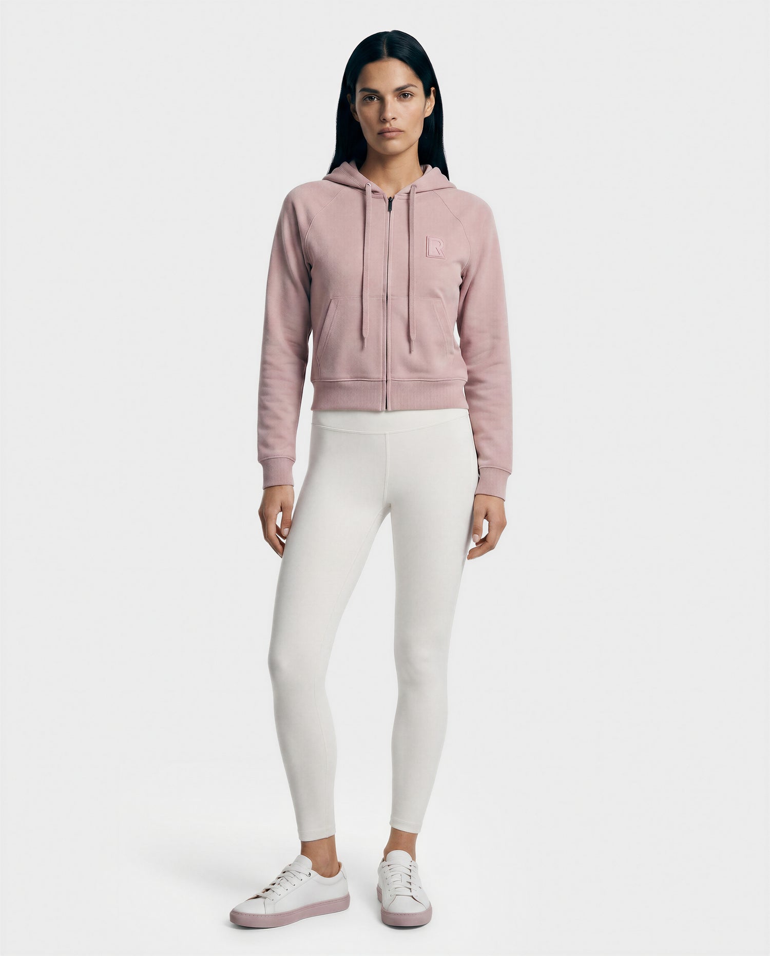 A woman faces forward against a white background, wearing the Rudsak SAMMY - L523014T top in soft pink, paired with white fitted leggings and pink-and-white sneakers.