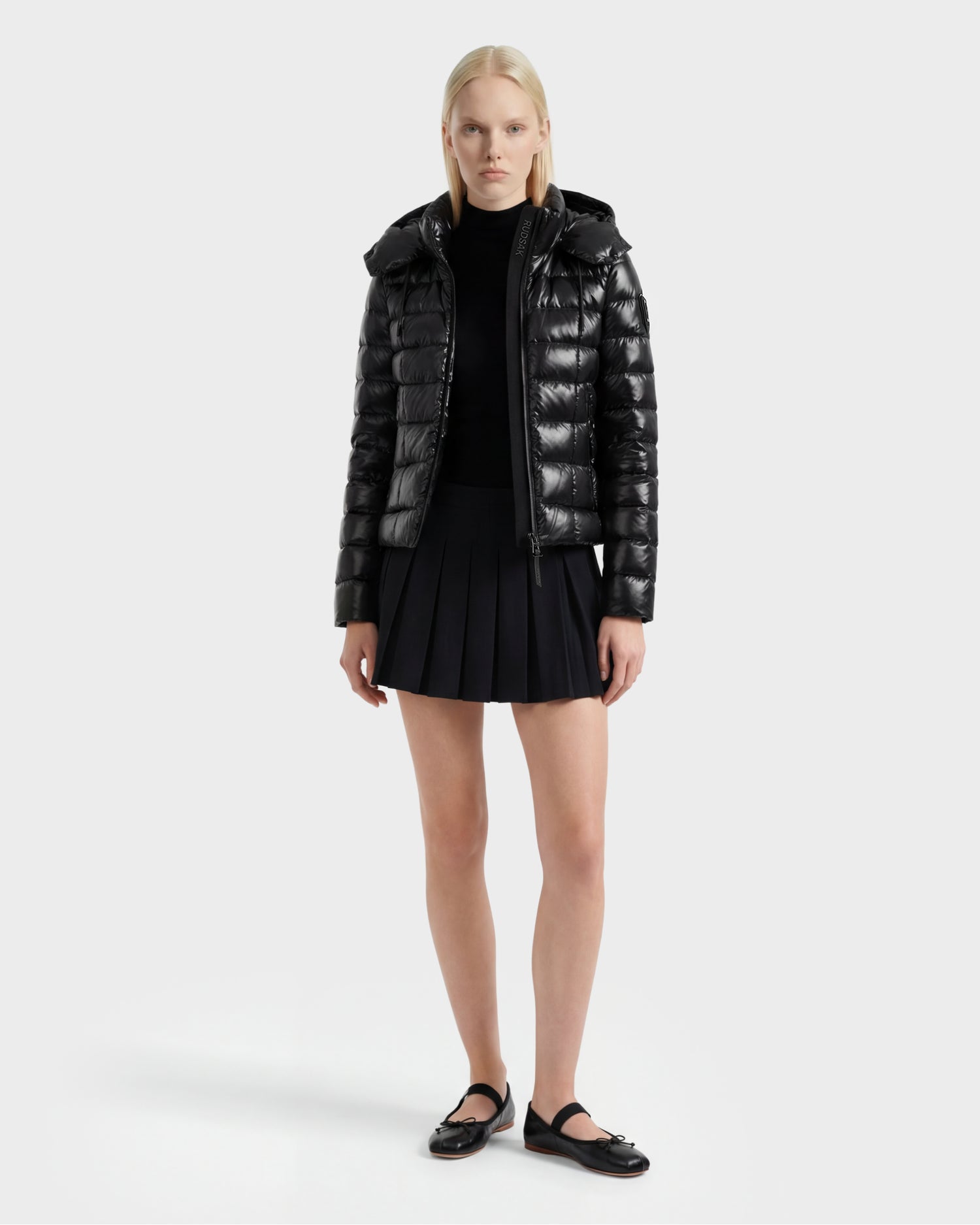 A woman with straight blonde hair and a neutral expression stands against a plain background wearing the Rudsak MELISSA C - 8125695 Black shiny puffer jacket, paired with a black pleated mini skirt, top, and flats.