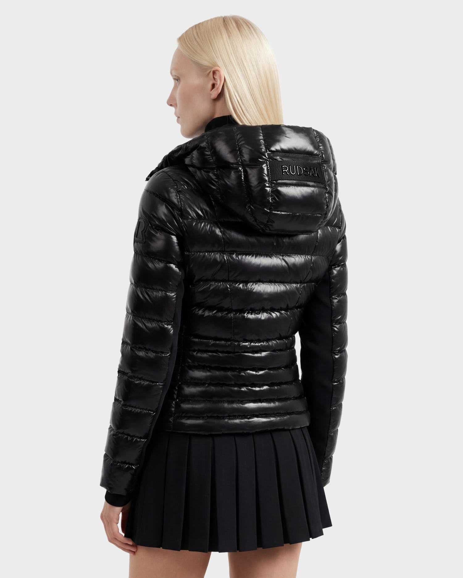 A person with straight, light blonde hair is seen from the back wearing a Rudsak MELISSA C - 8125695 Black women’s puffer jacket with a hood and a short black pleated skirt against a plain light gray background.