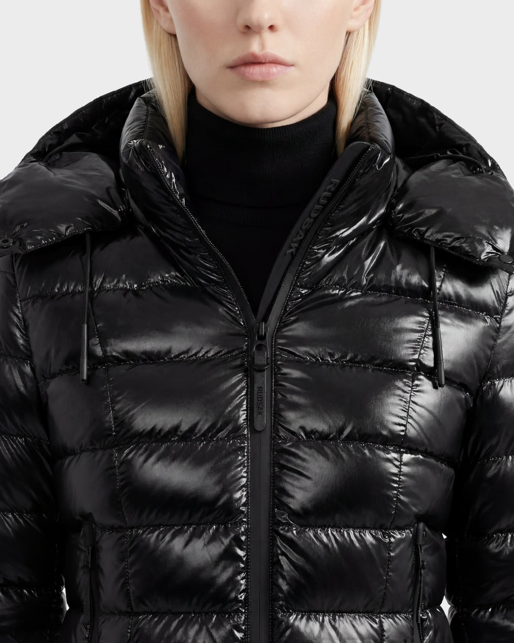 A person wearing the Rudsak MELISSA C - 8125695 Black women's puffer jacket with a hood and a black turtleneck sweater, shown from the chest up against a plain background.